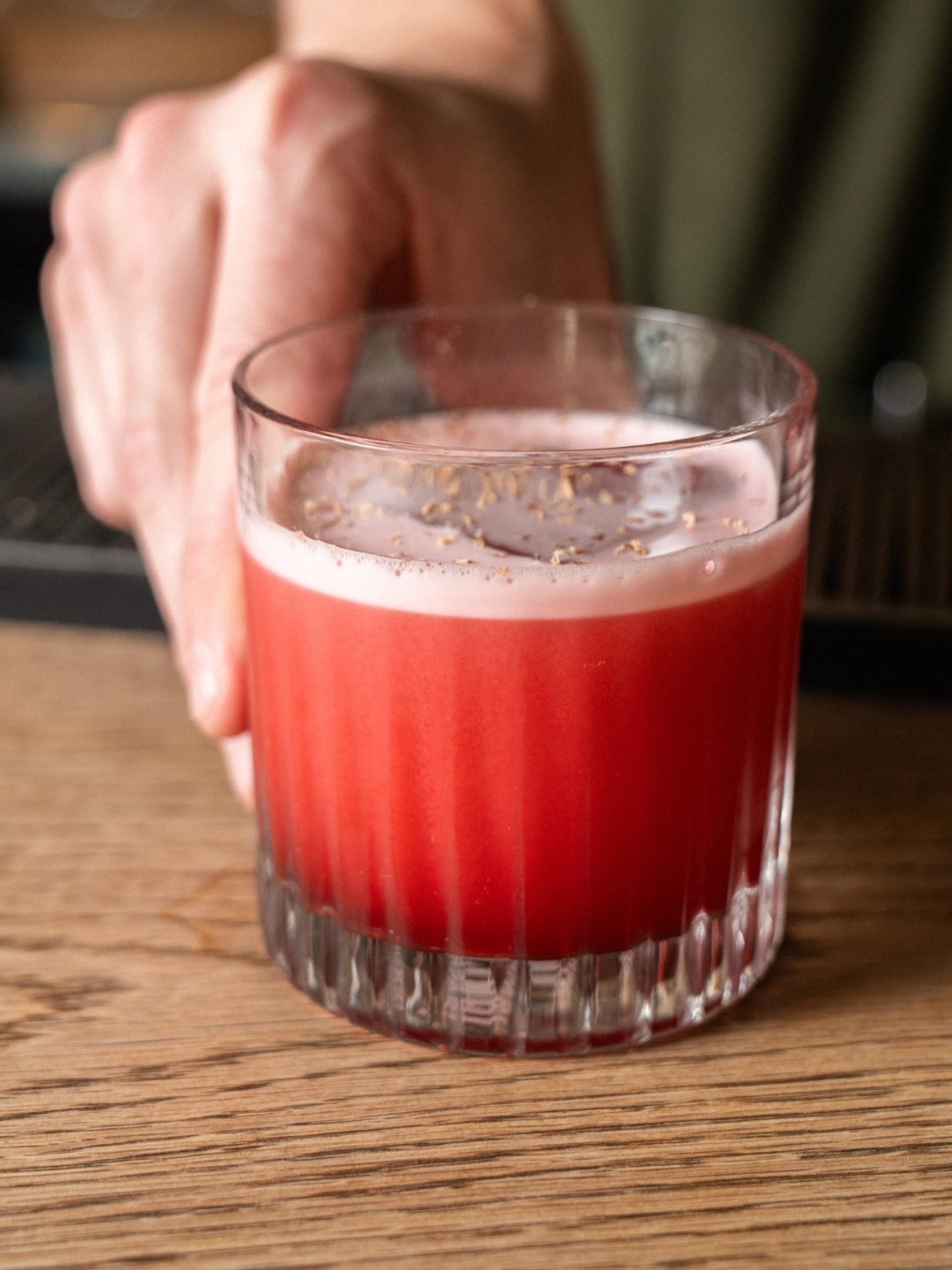 Hand holding a ribbed crystal glass with a deep red foamy cocktail on a wooden bar surface.