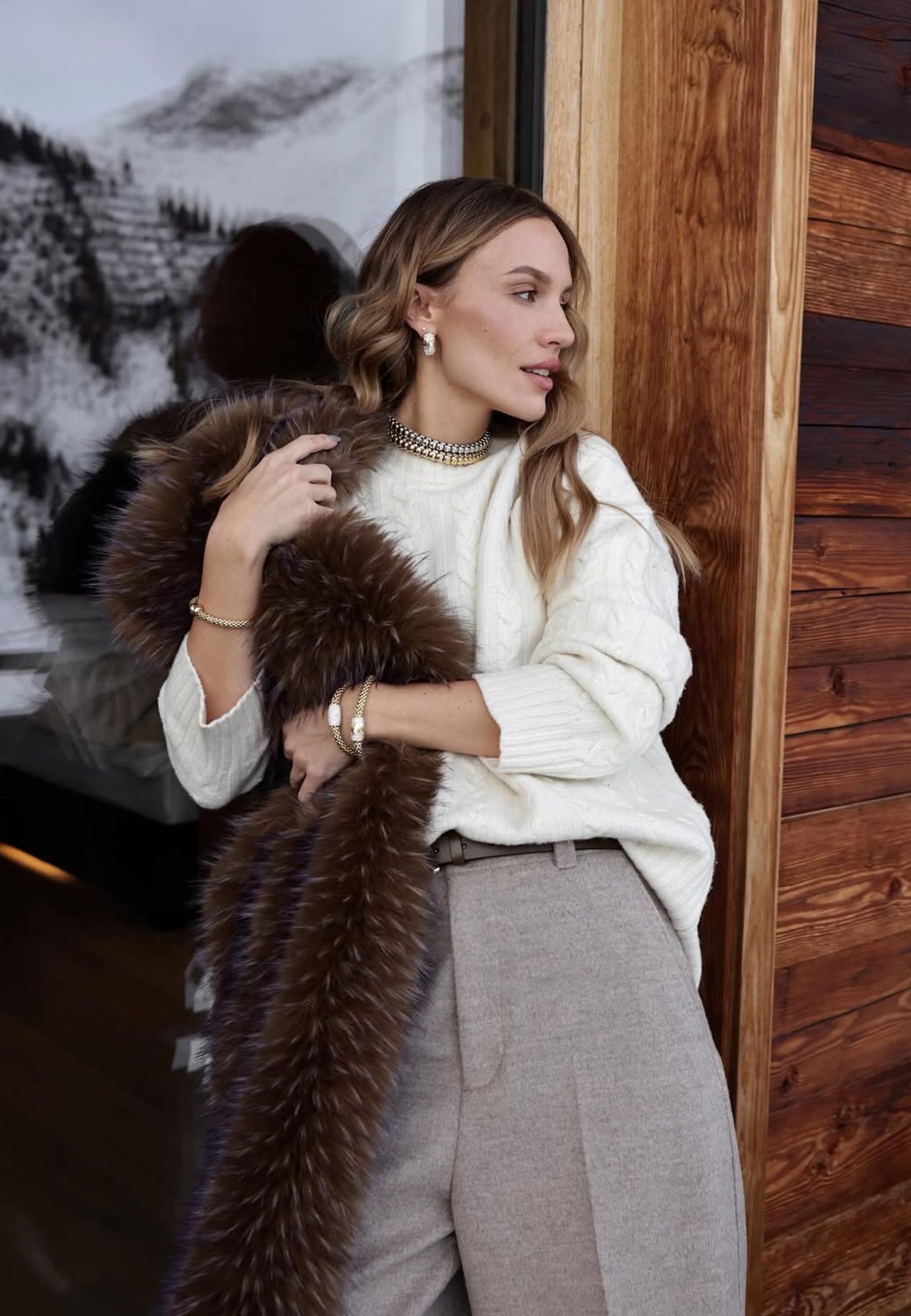 Woman in cable-knit sweater and fur stole wearing gold choker and bracelets in alpine interior.