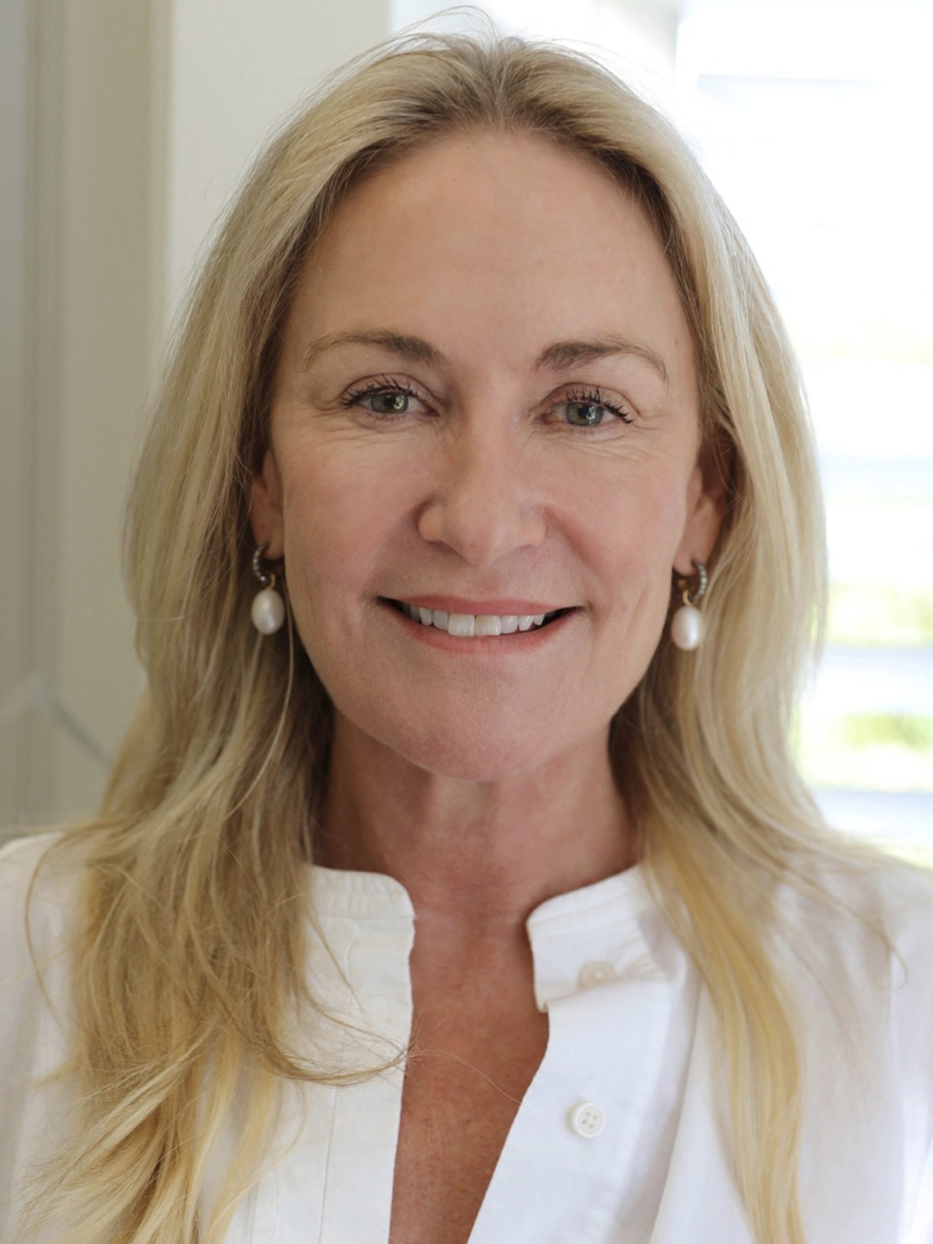 Smiling blonde woman in white linen shirt with pearl earrings, soft natural light background.