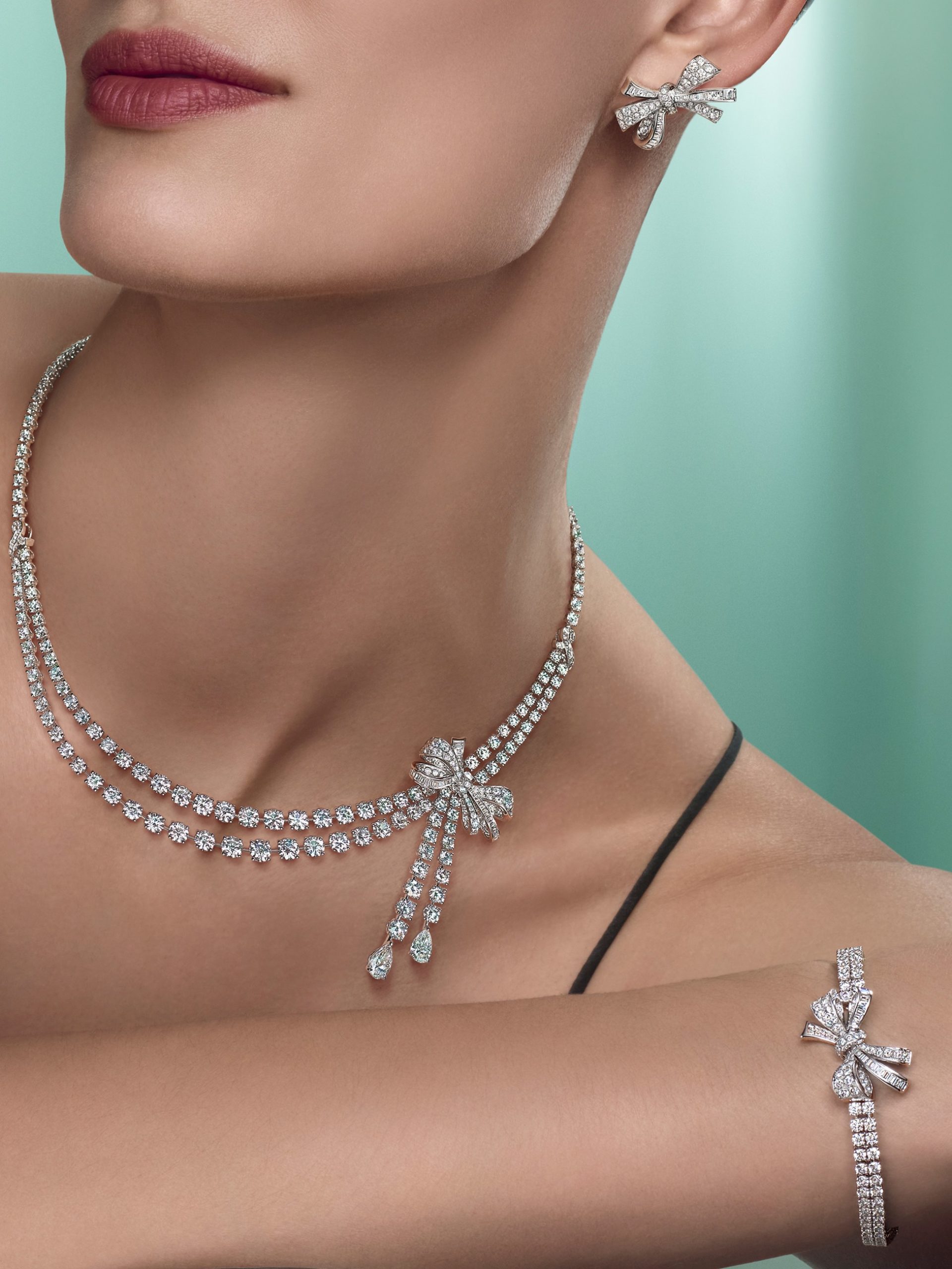 Model wearing Graff diamond bow necklace, earrings, and bracelet set against mint green background.