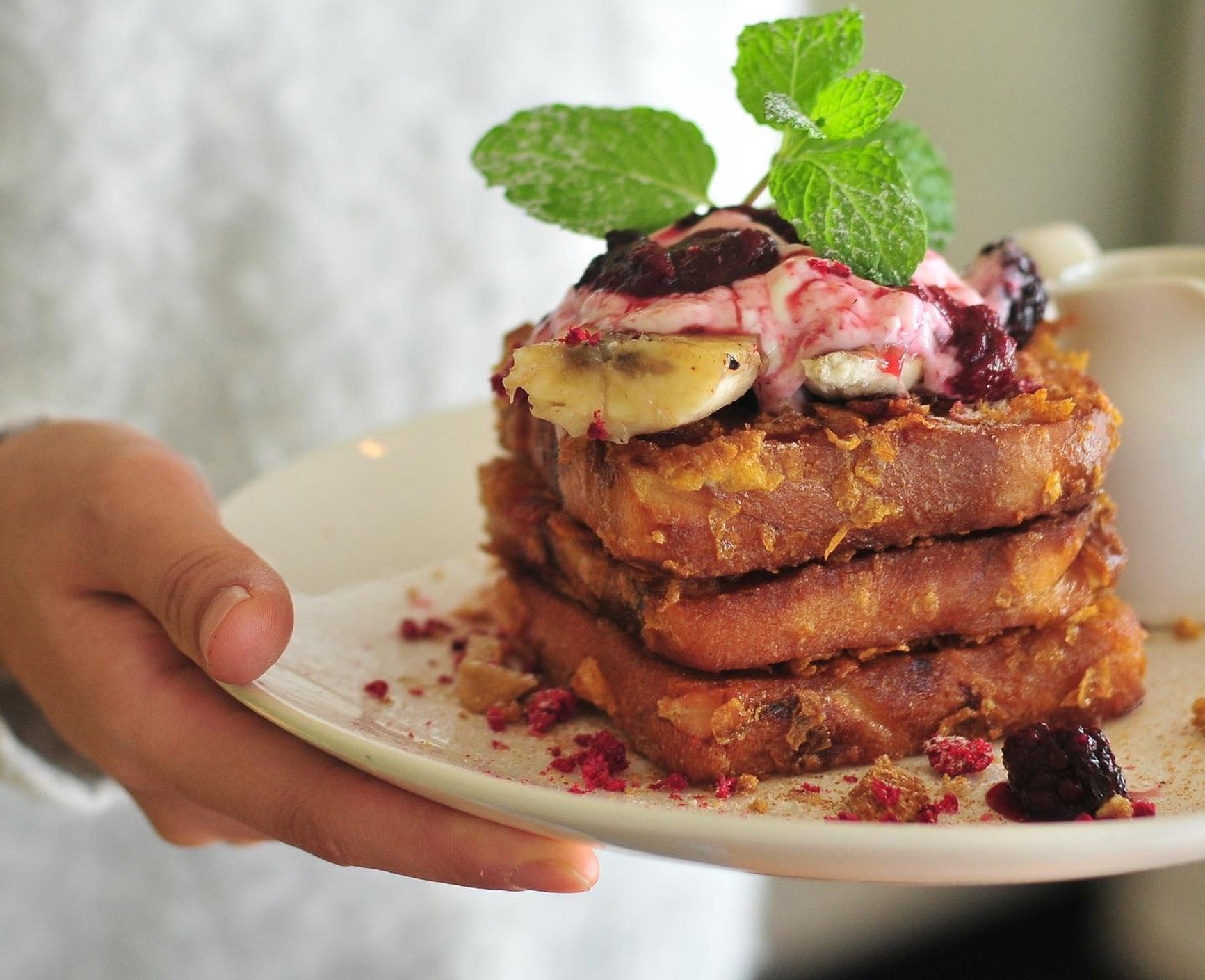 A hand holding a plate of stacked cornflake-crusted French toast topped with berries, banana, cream, and mint.