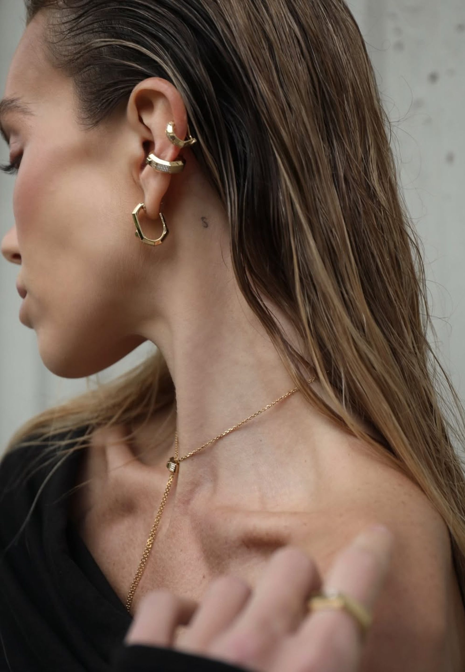 Woman in profile wearing stacked gold geometric ear cuffs, hoop earrings, and a gold lariat necklace.