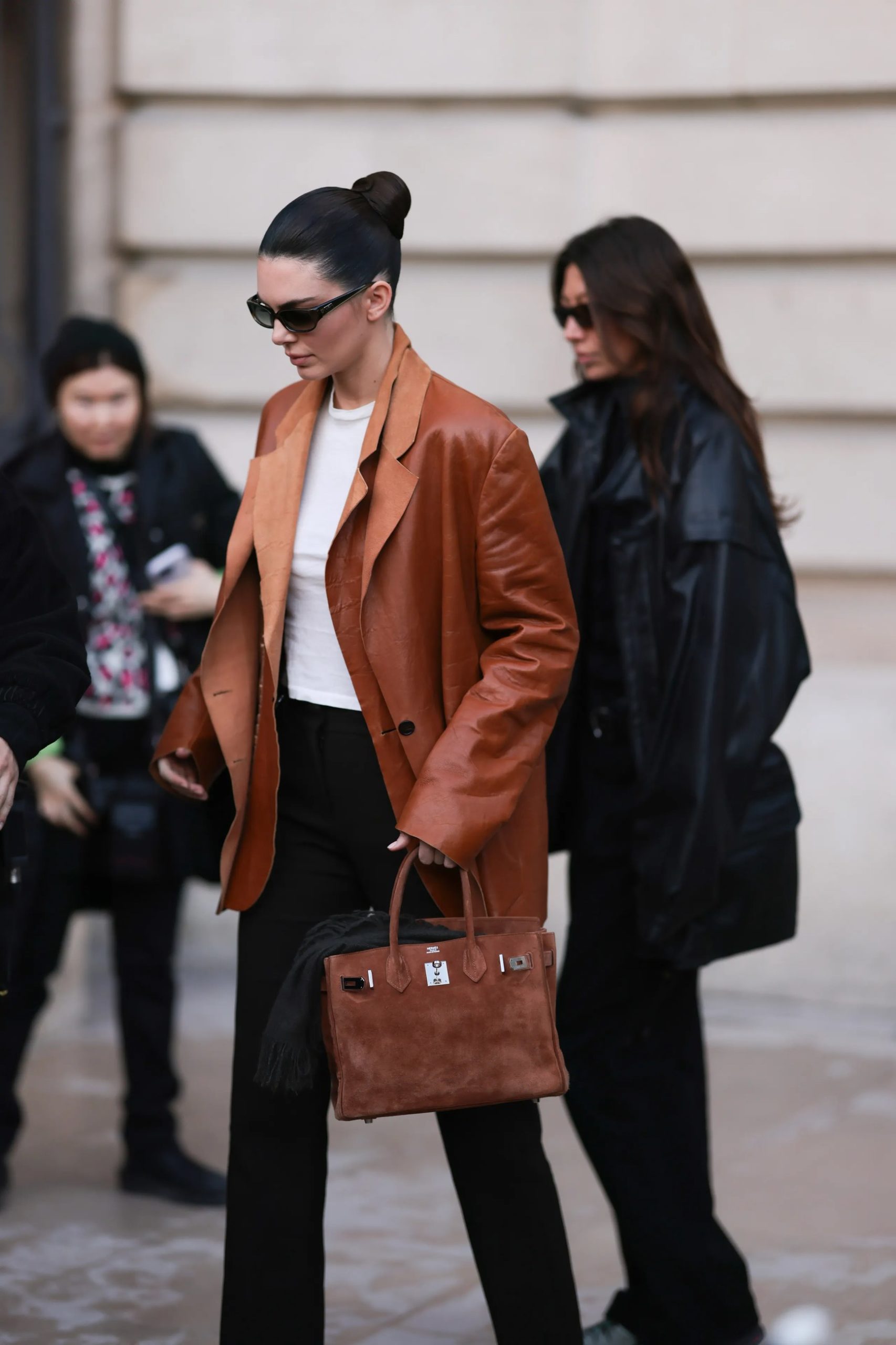 Woman in brown leather blazer carrying a tan suede Hermès Birkin bag on a city street.