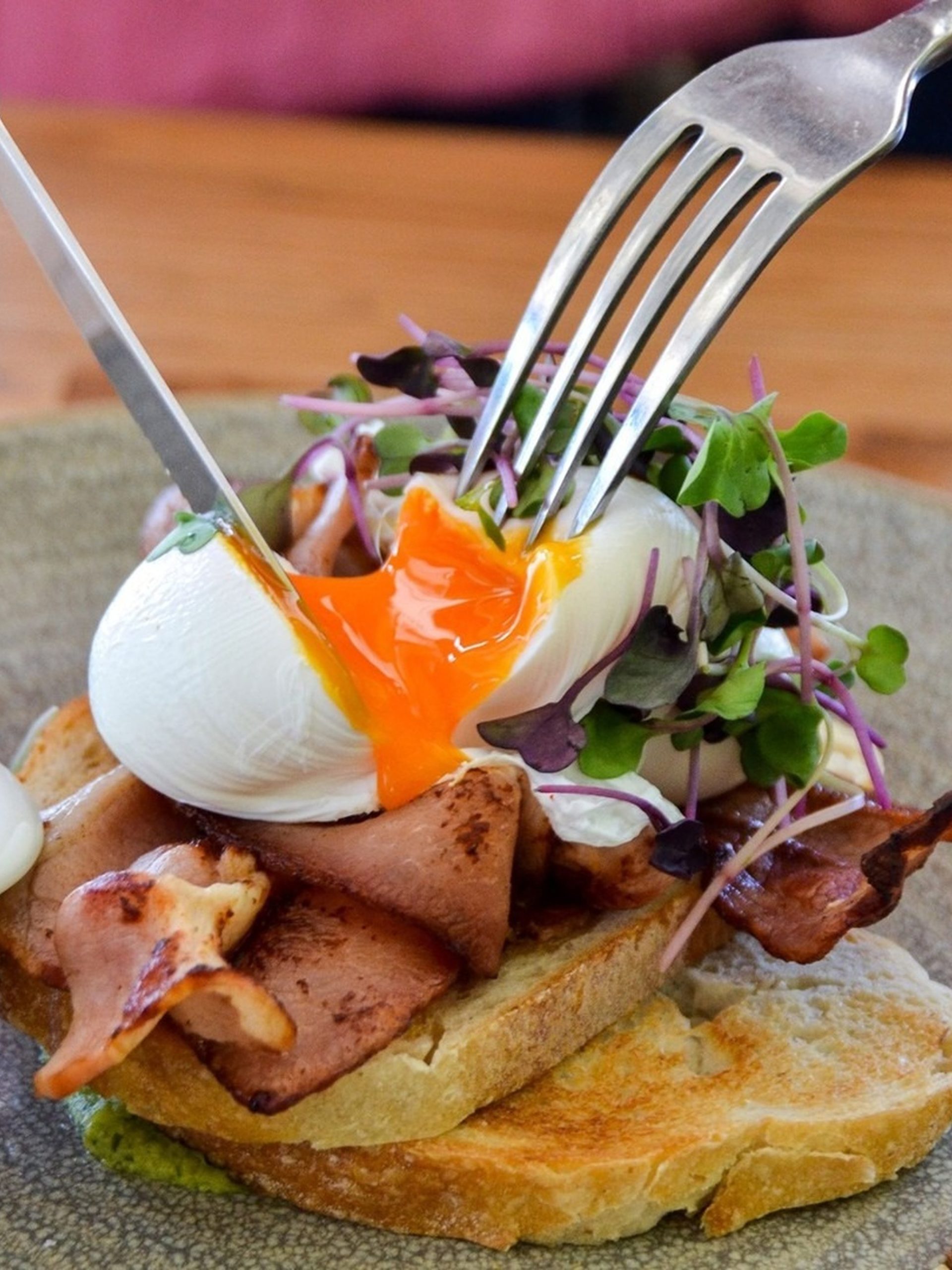 Poached egg cut open releasing golden yolk over bacon and sourdough toast with microgreens.