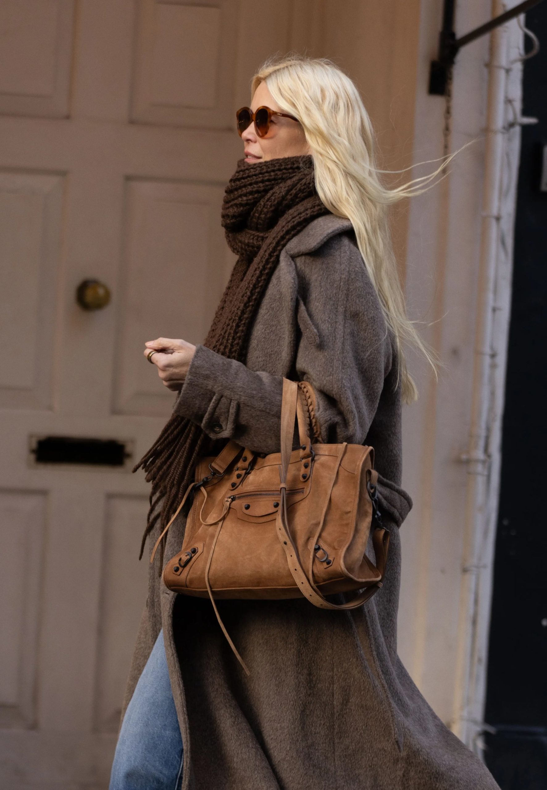 Blonde woman in long taupe coat and brown scarf carrying a tan Balenciaga bag on a city street.
