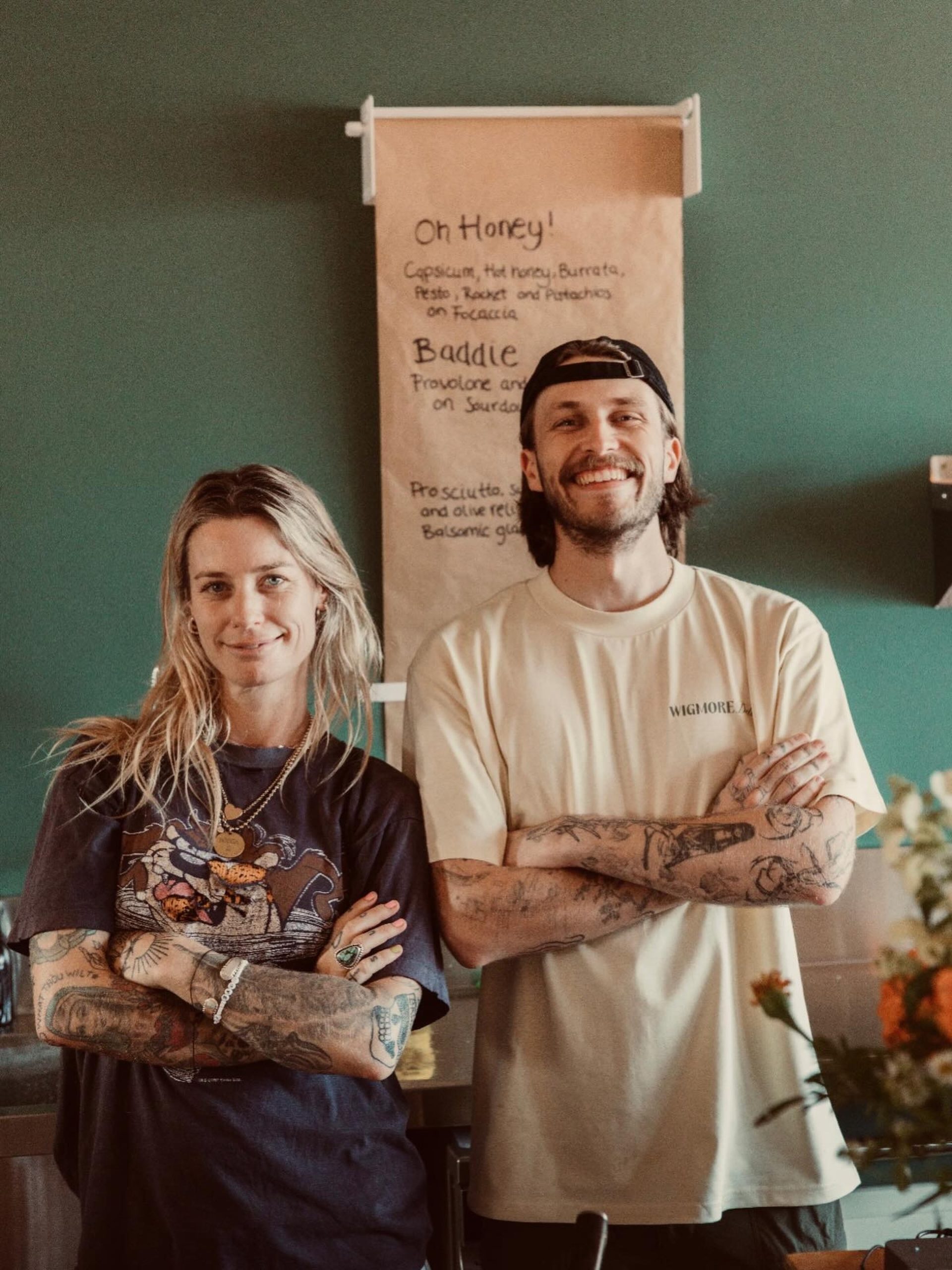Two tattooed people stand arms crossed, smiling in front of a handwritten café menu on a teal wall.