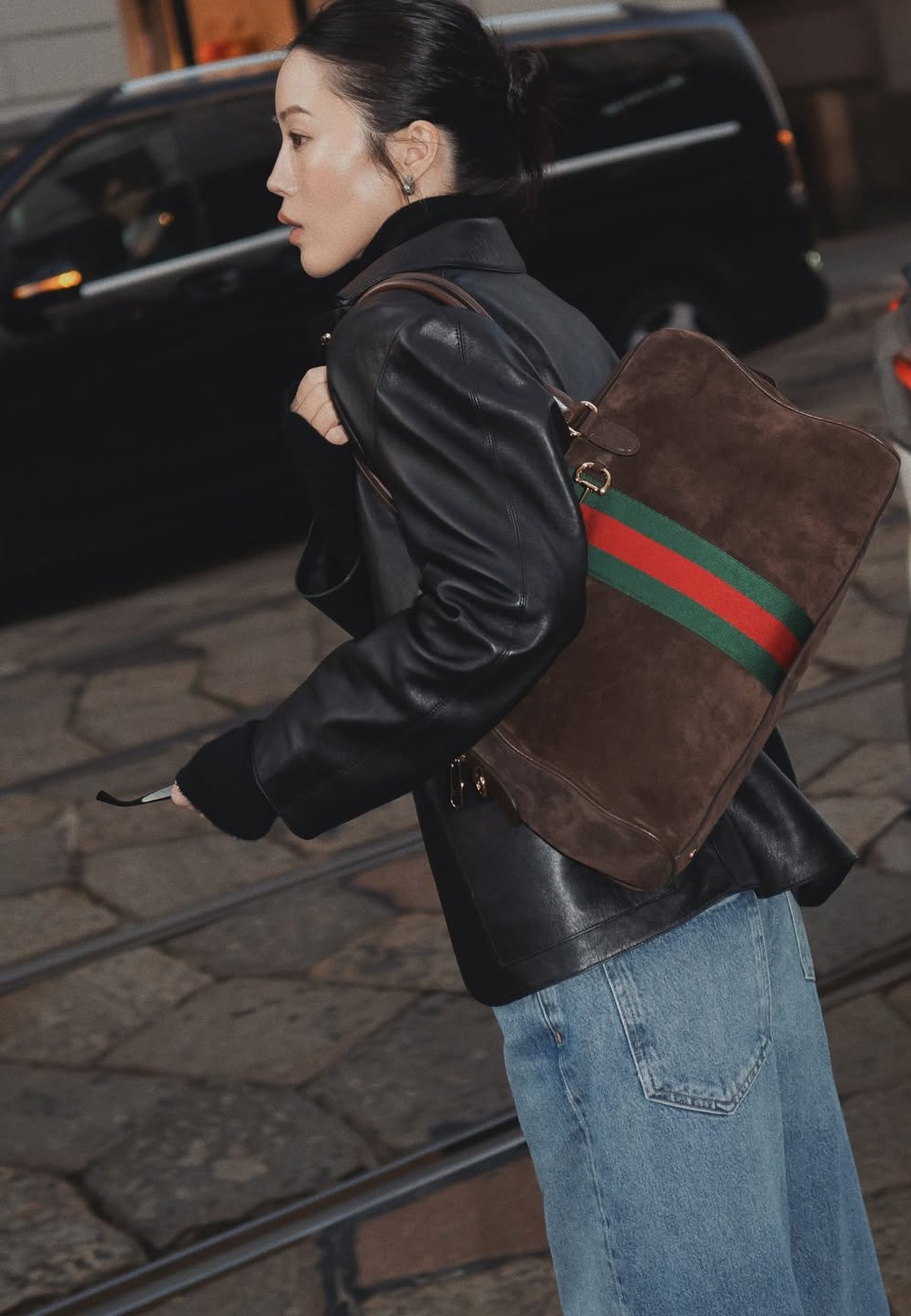 Woman in black leather jacket carrying a brown suede Gucci Web stripe backpack on cobblestone street.