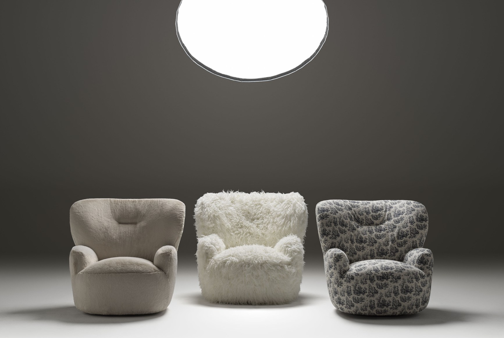 Three plush armchairs in taupe, white shag, and toile fabric under dramatic studio lighting.