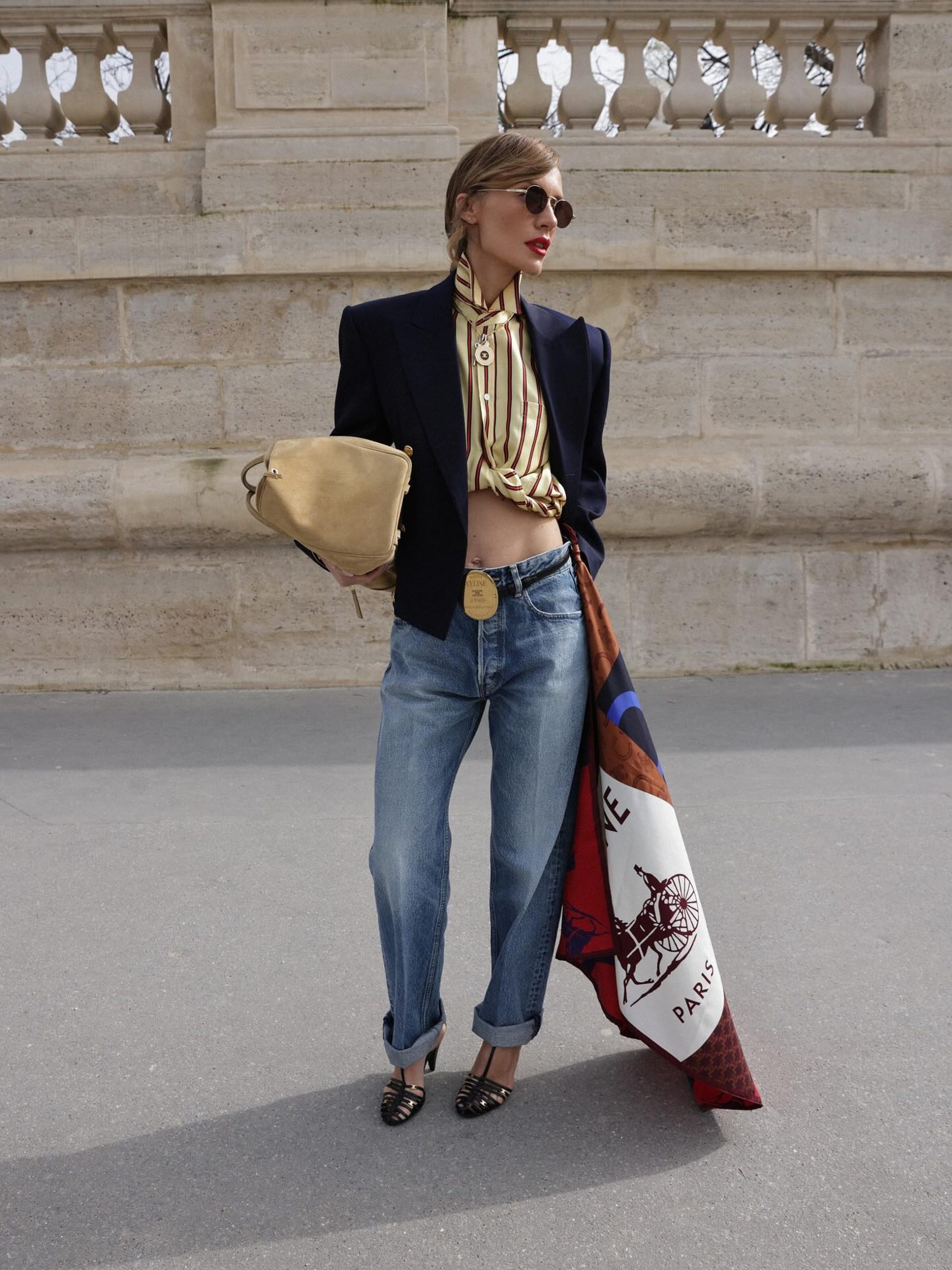 Woman in navy blazer, striped shirt, cuffed jeans and heels holding Hermès scarf on Paris street.