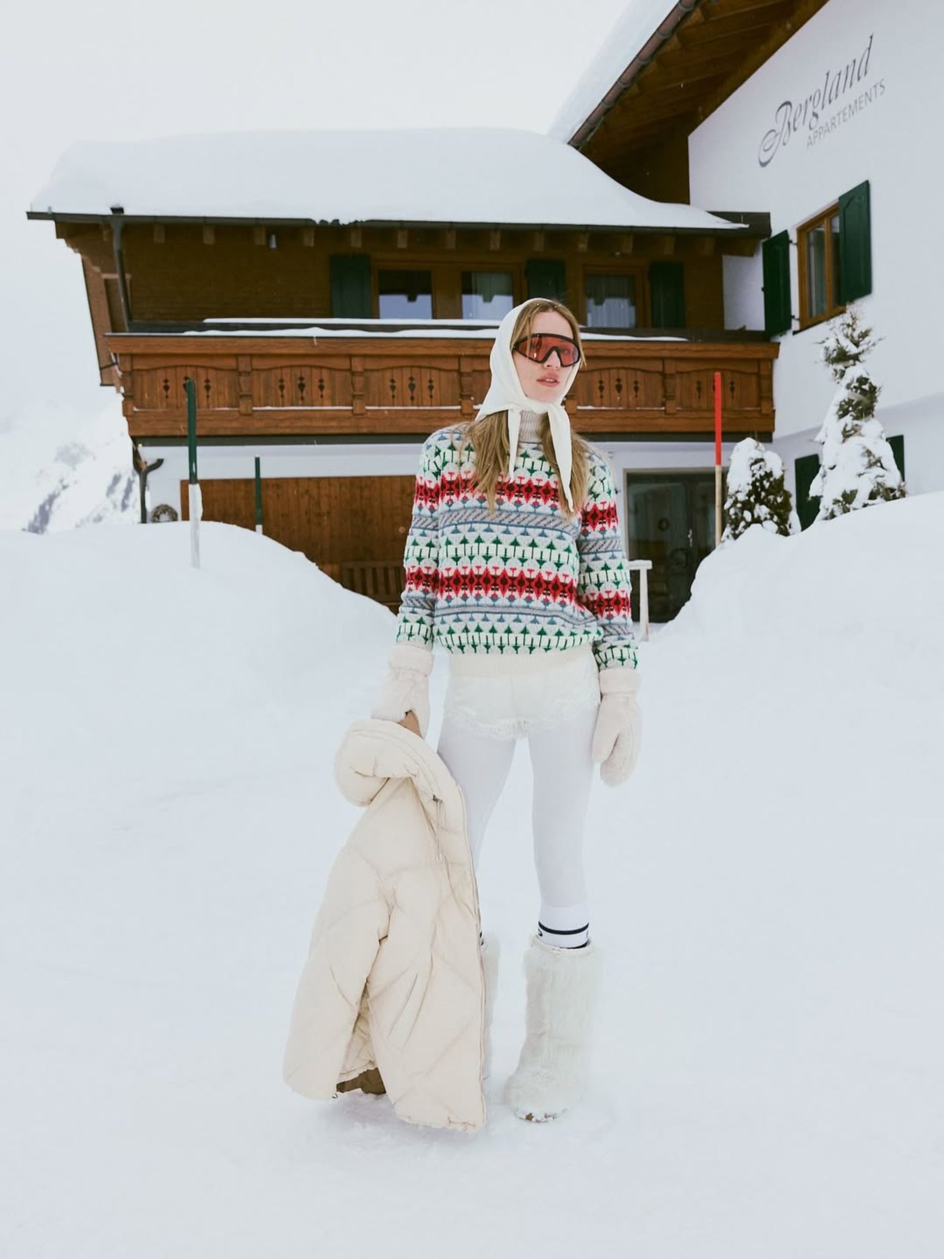 Woman in Fair Isle sweater and ski goggles standing in snow outside an Alpine chalet.