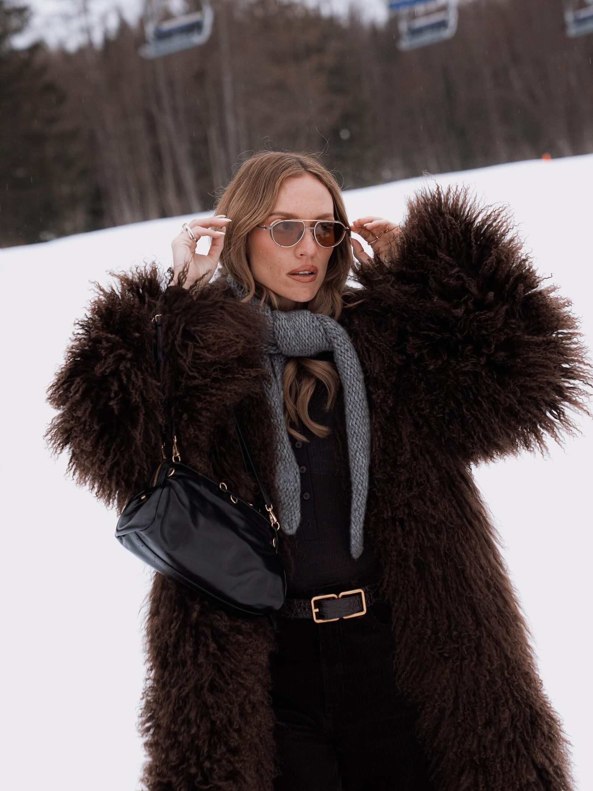 Woman in oversized brown fur coat and aviator sunglasses posing on a snowy alpine slope.