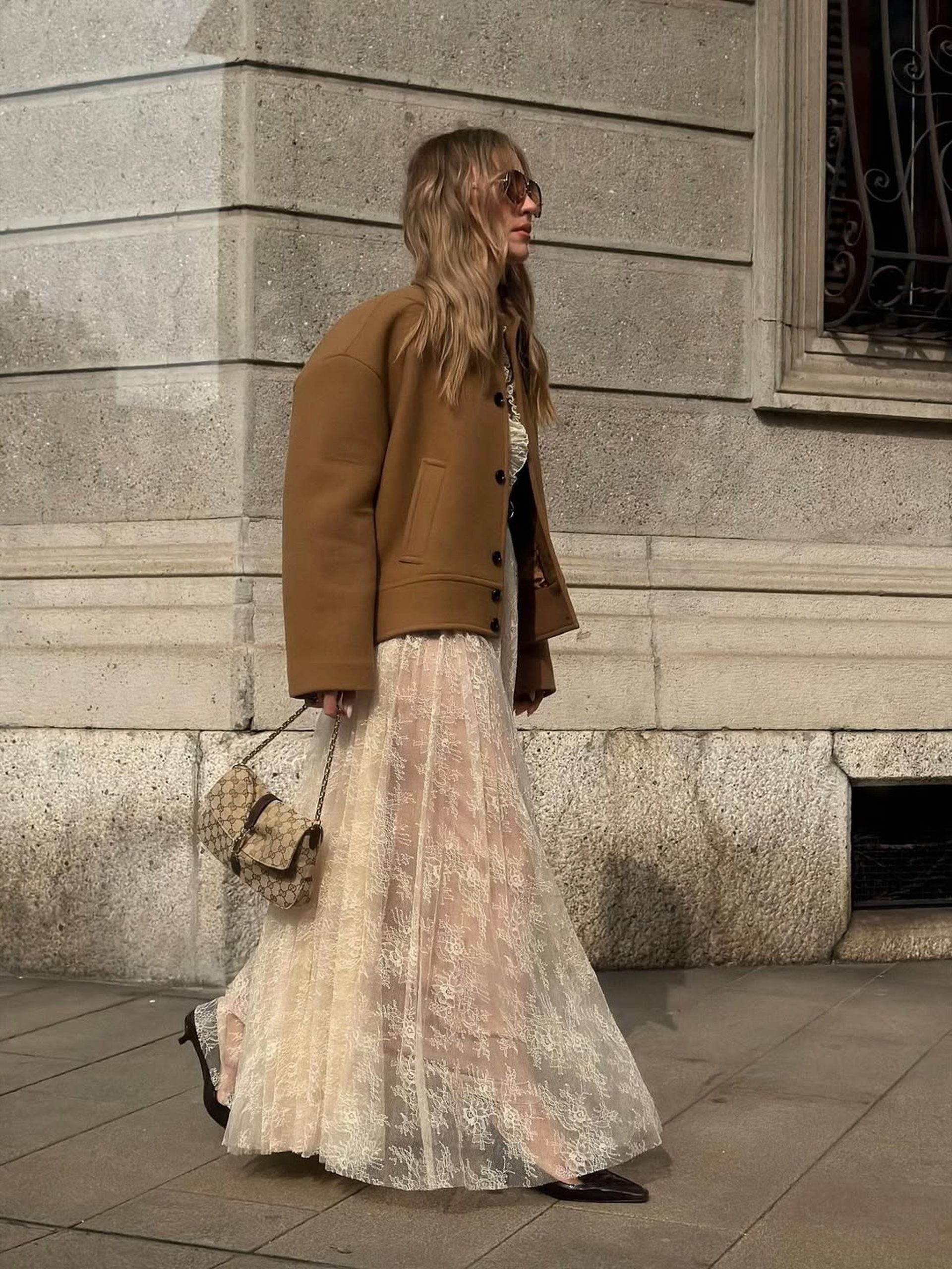 Blonde woman in camel jacket and ivory lace skirt walking along a European stone street.
