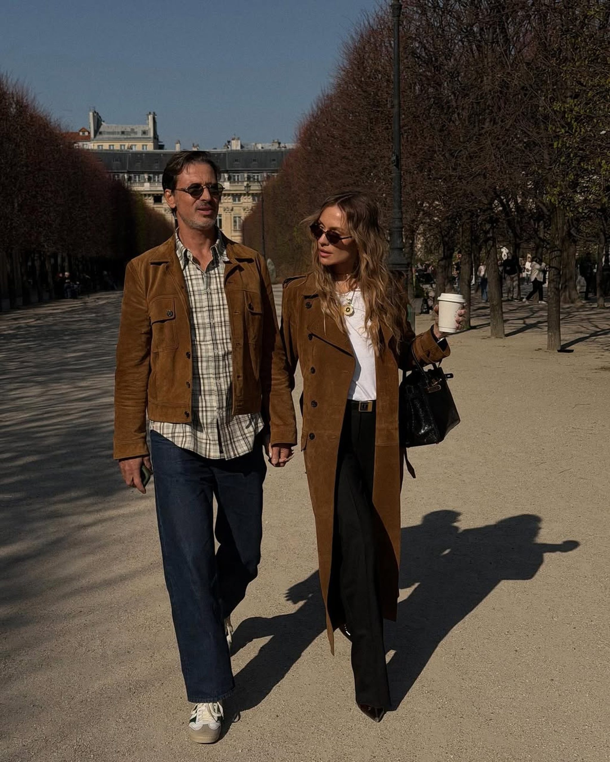 A couple walks through a Parisian garden in matching brown suede coats on a sunny day.