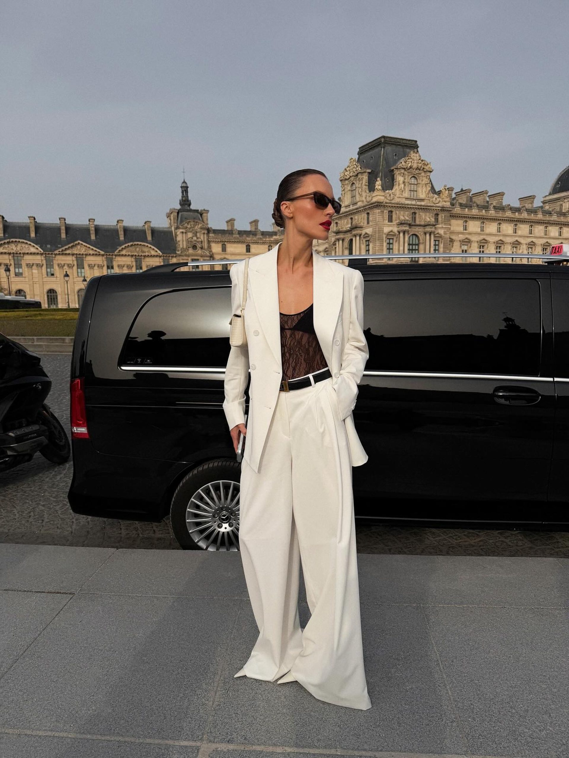 Woman in white tailored suit with sheer lace top standing before the Louvre, Paris.