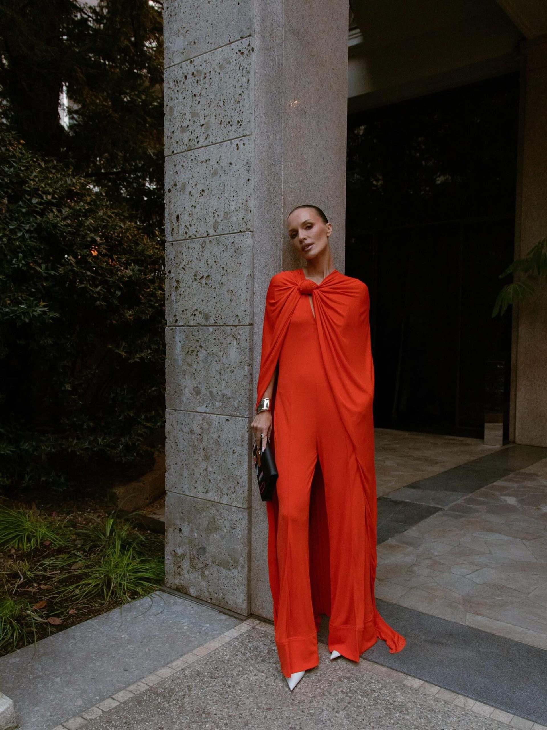 Woman in red cape jumpsuit leaning against stone column at a modernist building entrance.