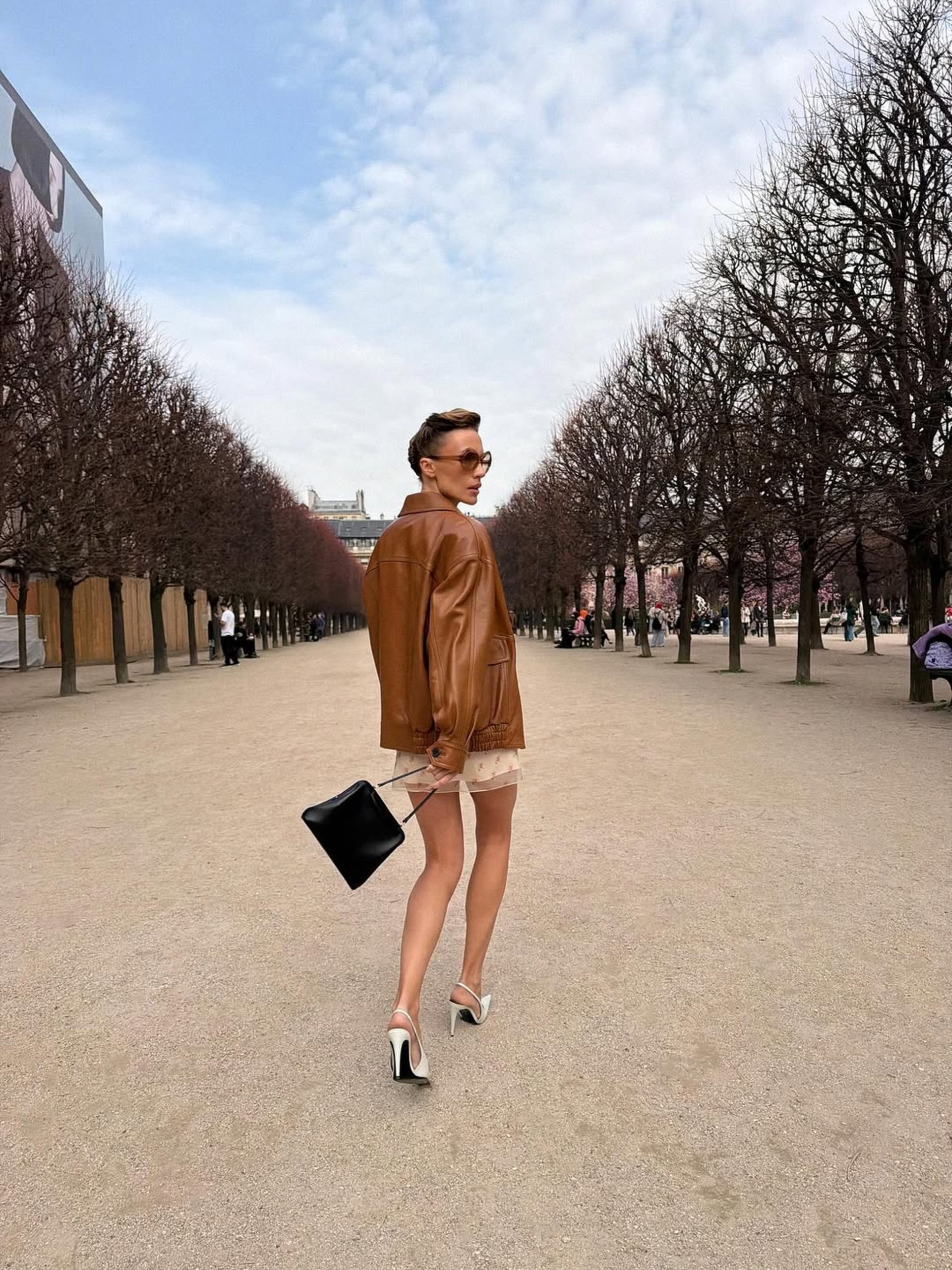 Woman in tan leather jacket and floral mini dress walking through a Parisian tree-lined promenade.