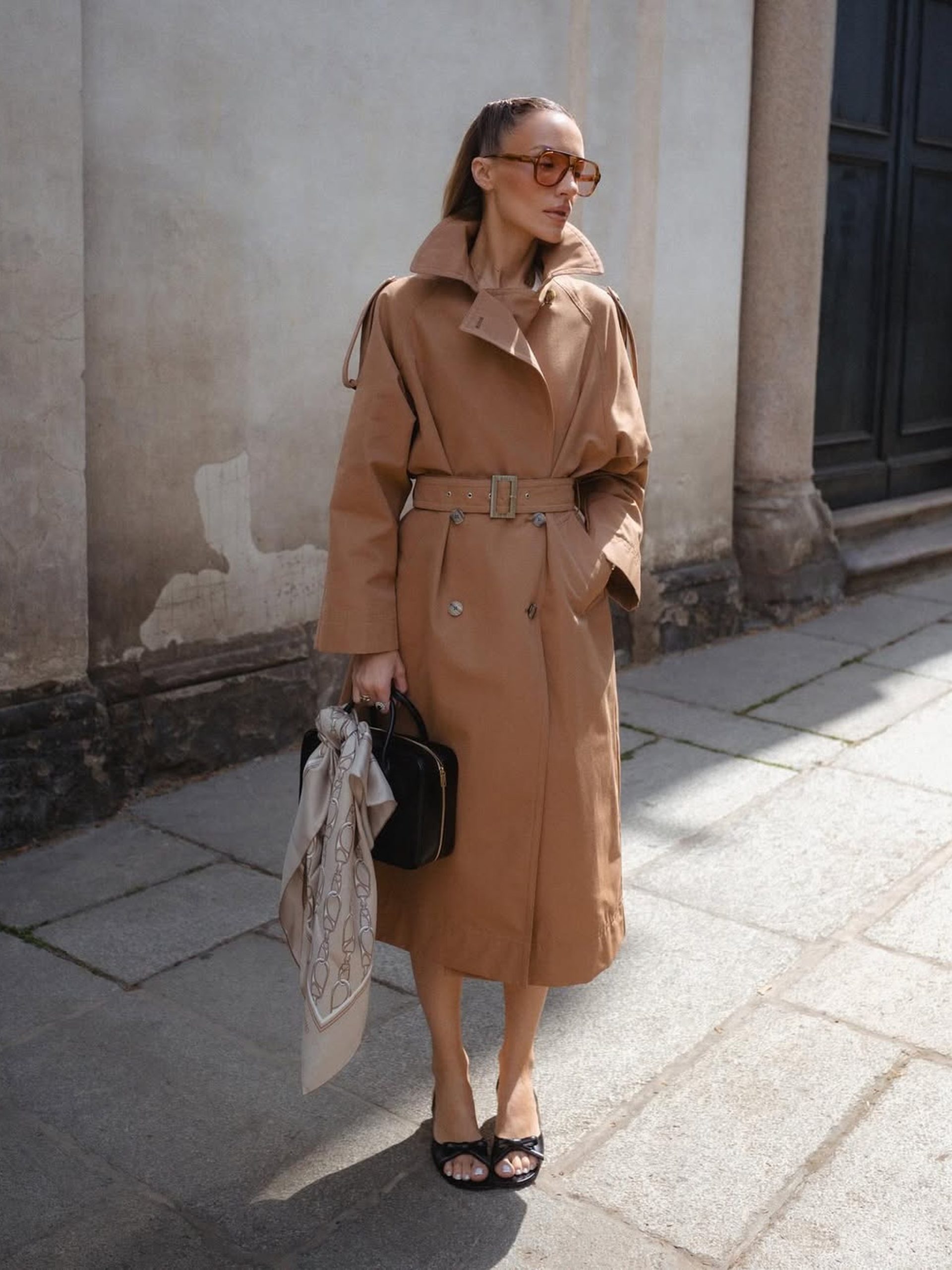 Woman in belted camel trench coat holding black handbag and silk scarf on cobblestone street.