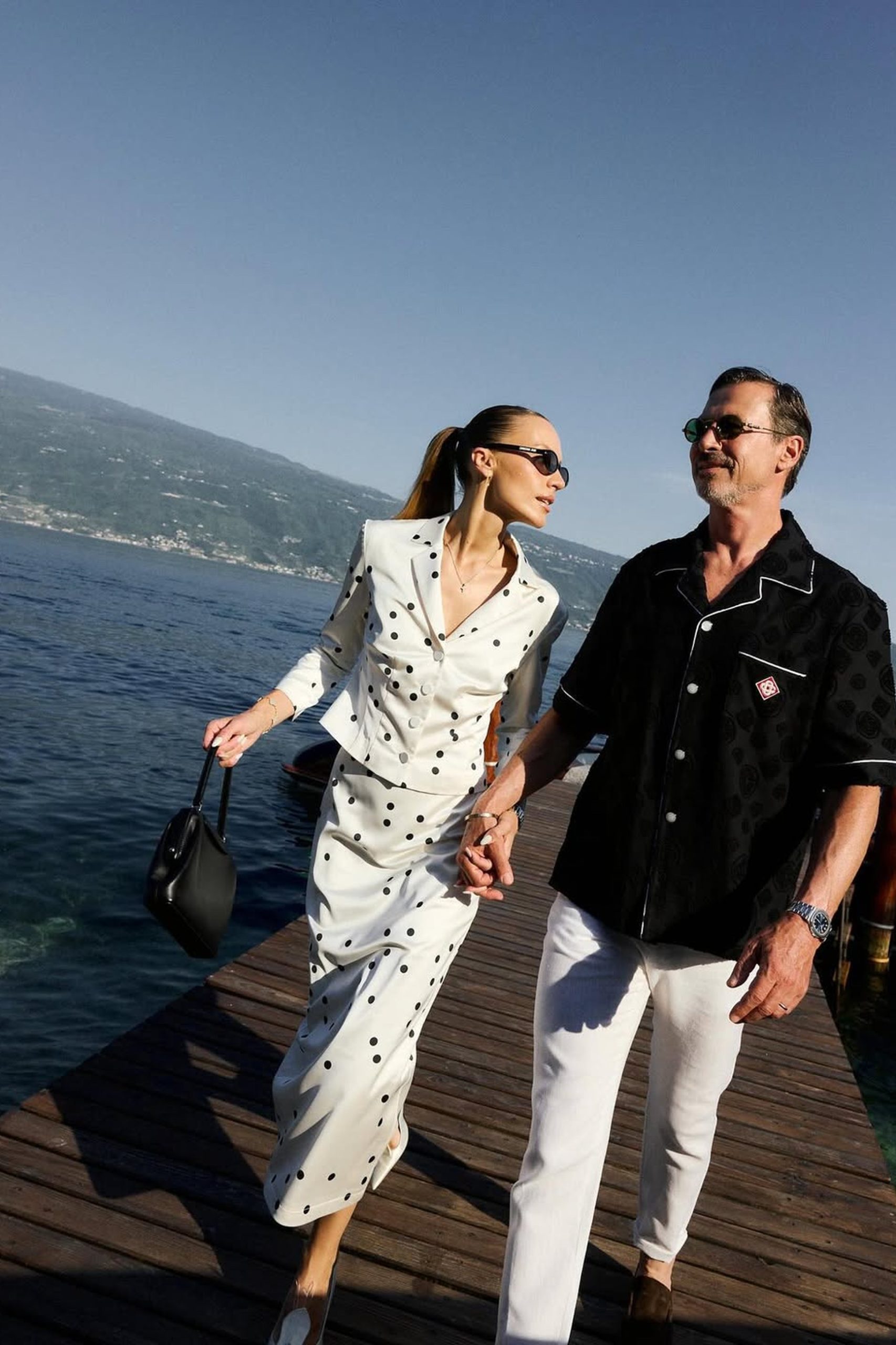 Stylish couple walking hand-in-hand along a wooden dock beside a blue European lake.
