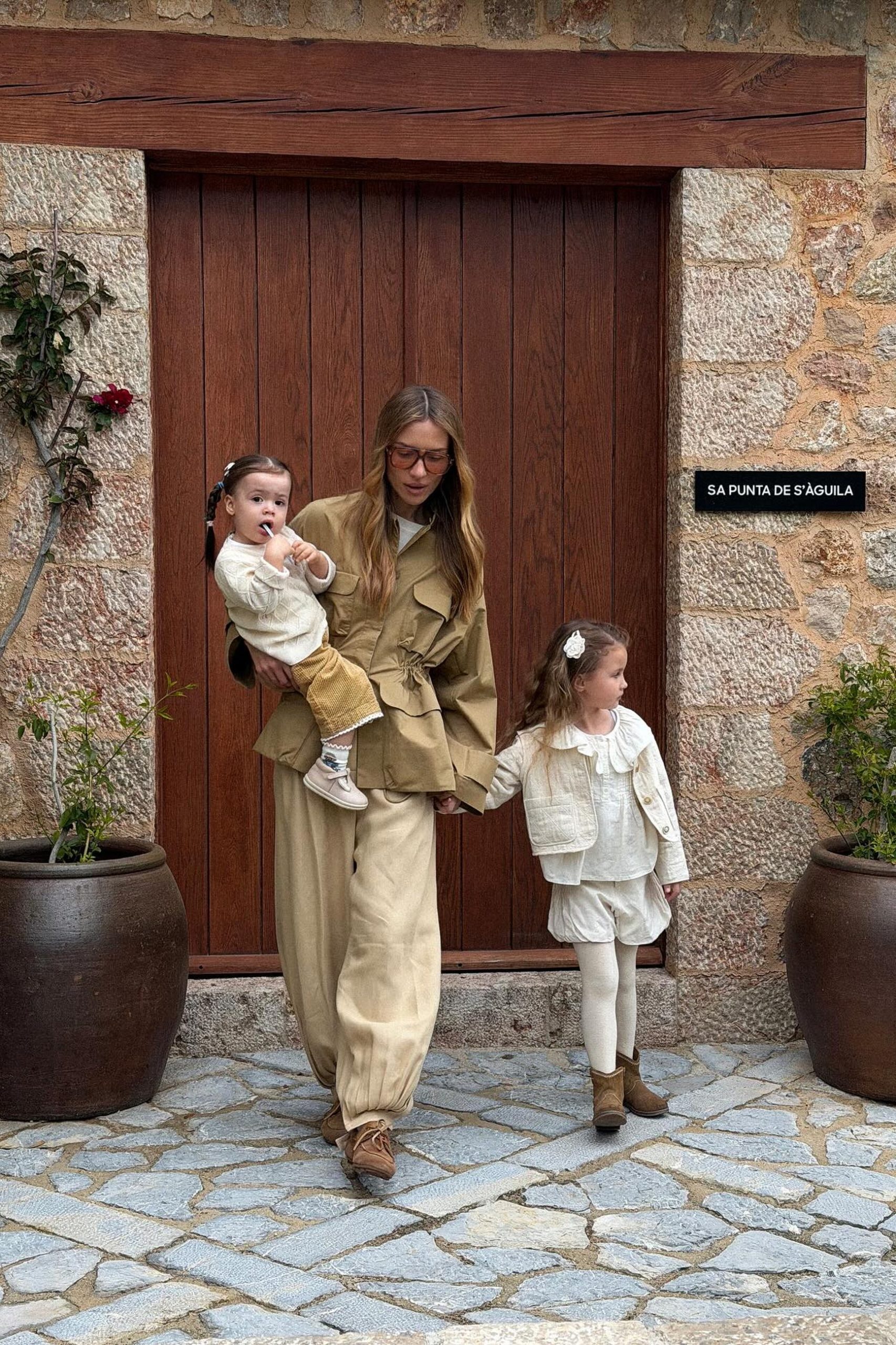 Woman in olive utility outfit holds toddler and walks with young daughter outside rustic stone building.