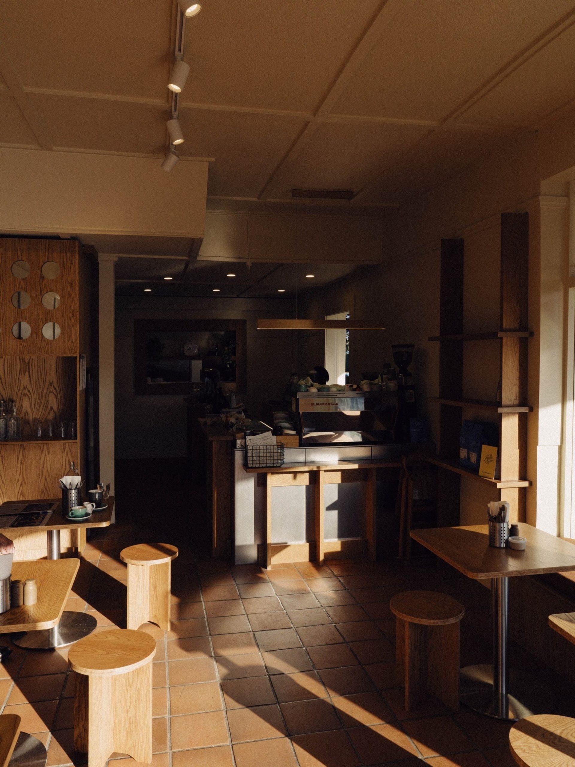 Sunlit café interior with light oak furniture, terracotta tile floors, and espresso bar in background.
