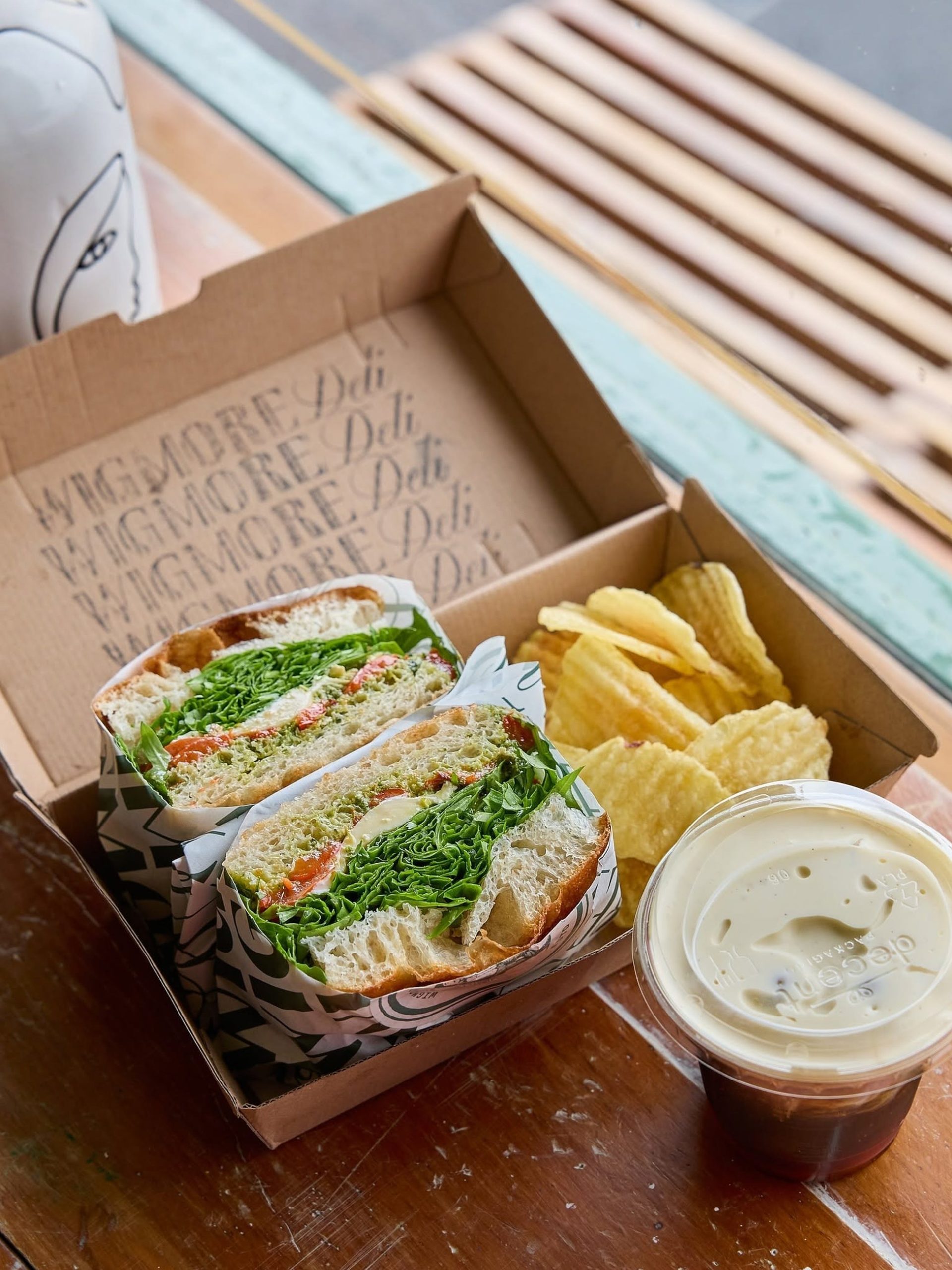 Open Wigmore Deli lunch box with ciabatta sandwich, ridged crisps, and dipping sauce on wooden bench.