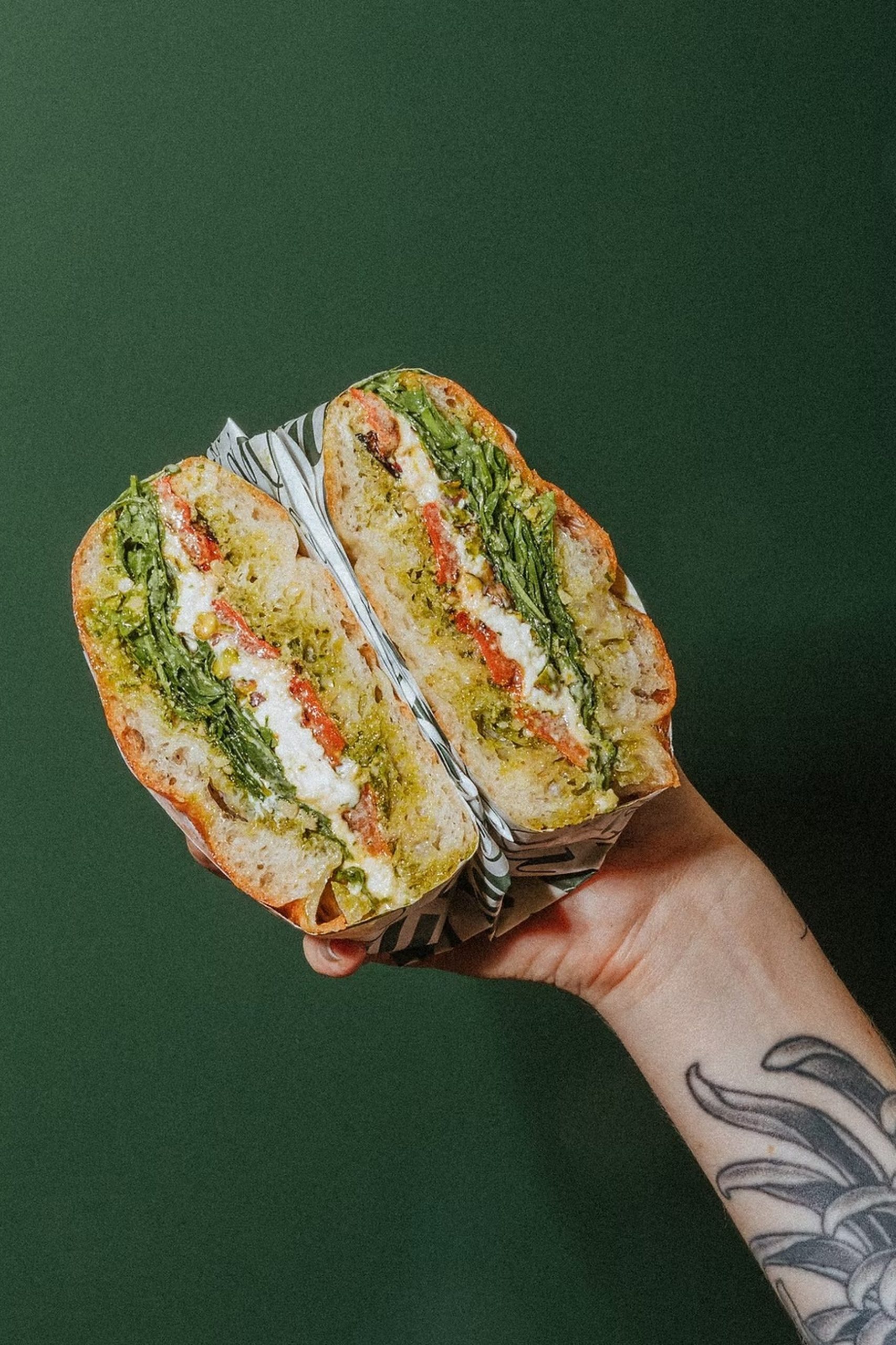 Tattooed hand holding a sliced sandwich with pesto, roasted peppers, greens, and white cheese on sourdough.