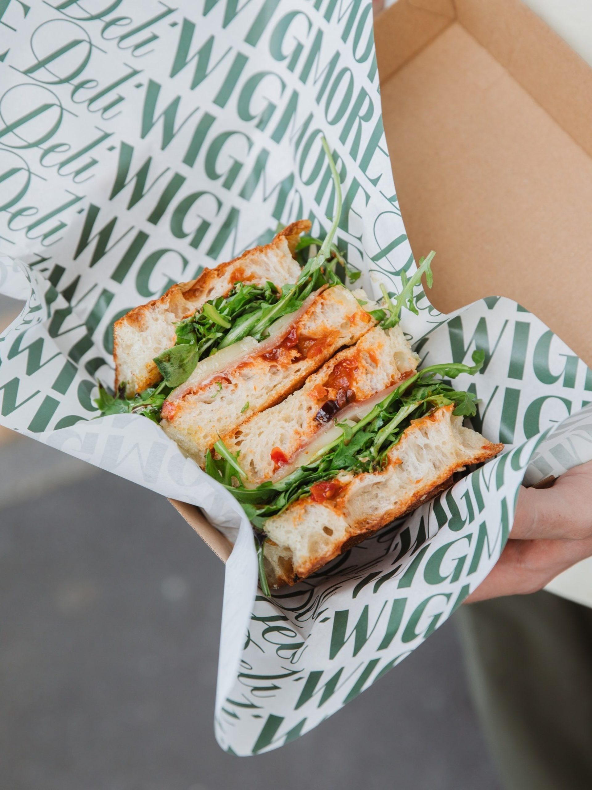 Hand holding an open box with a sourdough sandwich filled with ham, cheese, and rocket, wrapped in Wigmore Deli branded paper.