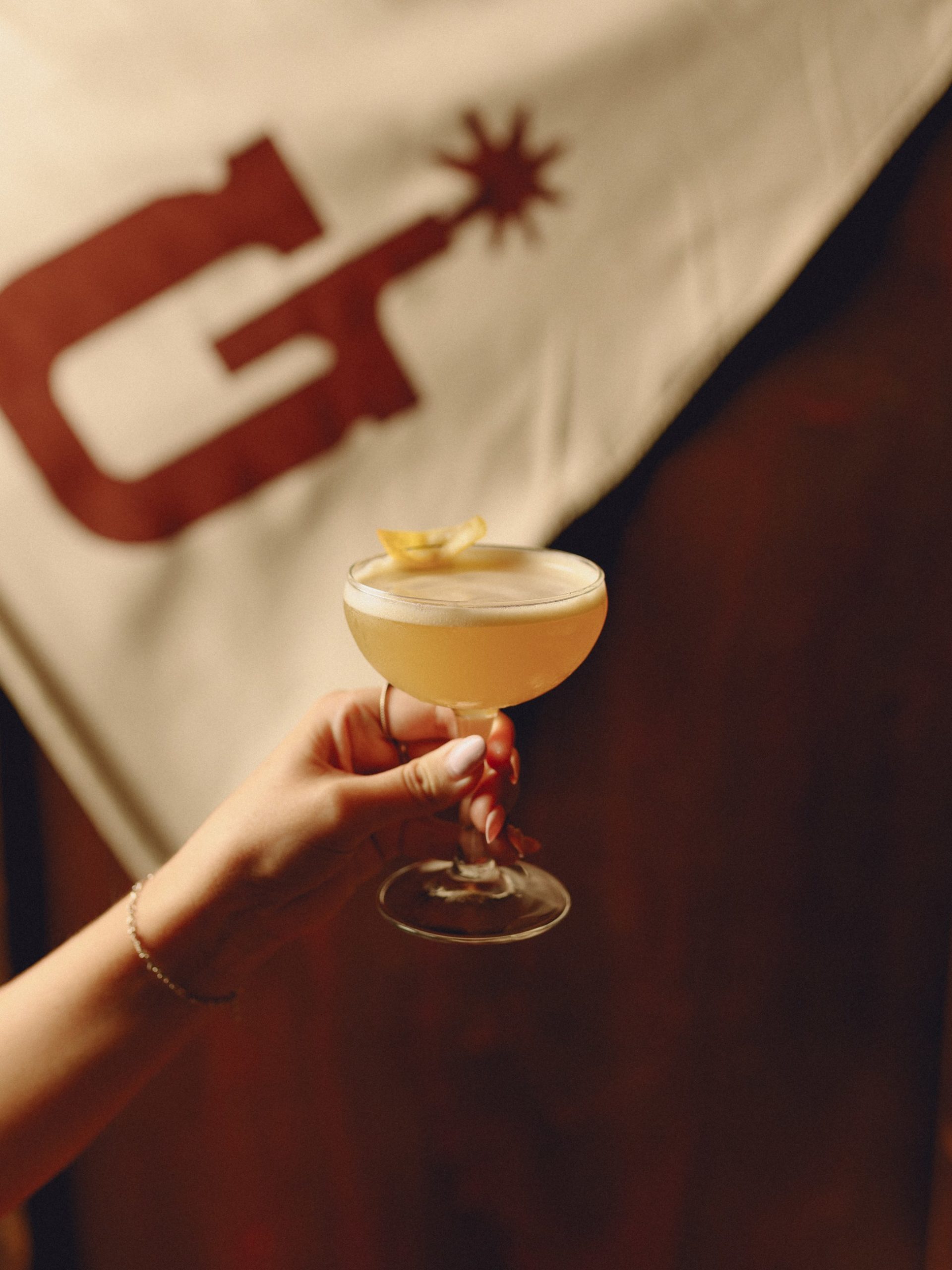 A hand holding a pale golden cocktail in a coupe glass with a citrus twist garnish.