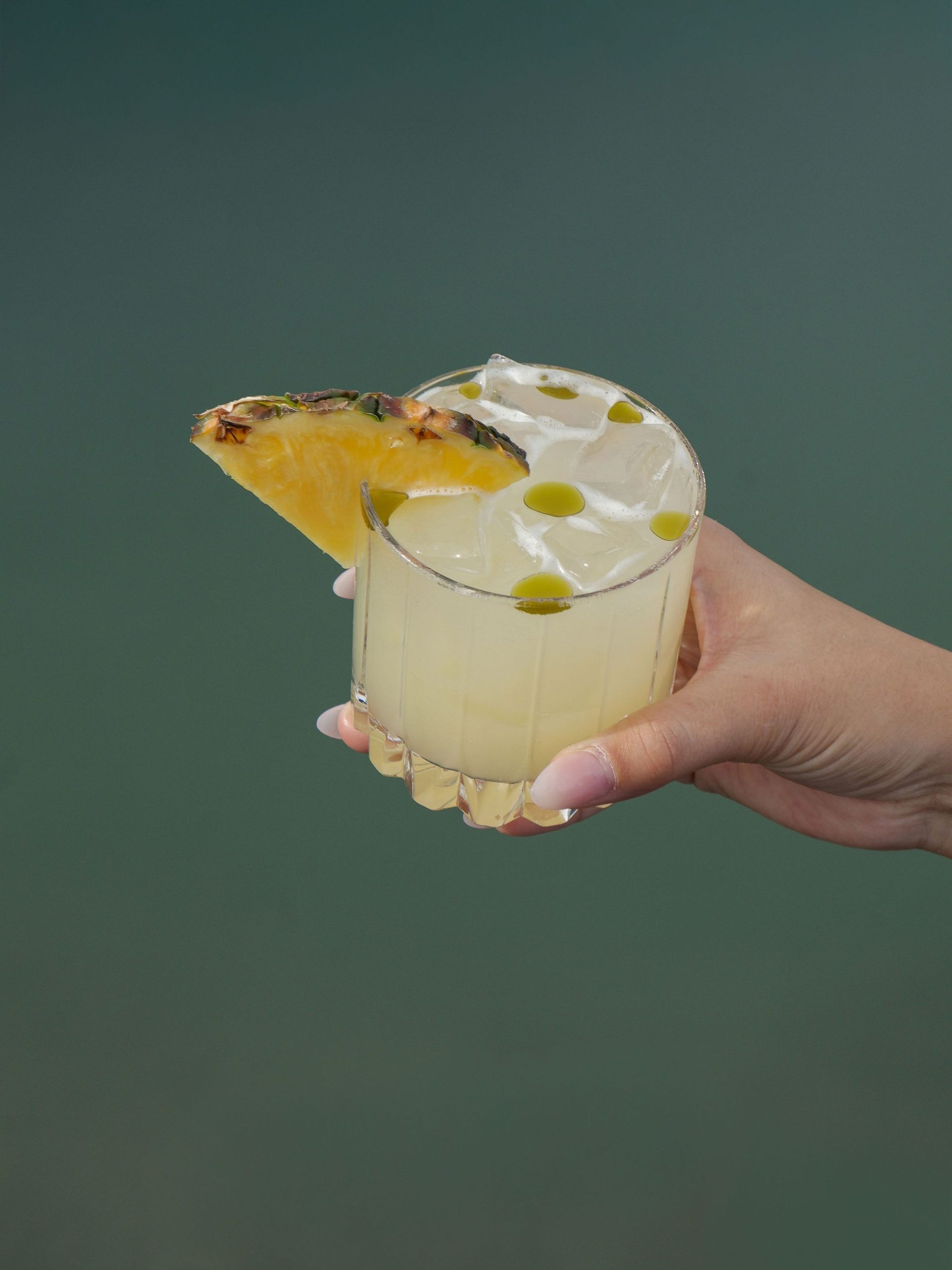 Hand holding a pineapple-garnished pale yellow cocktail in a faceted glass against a teal background.