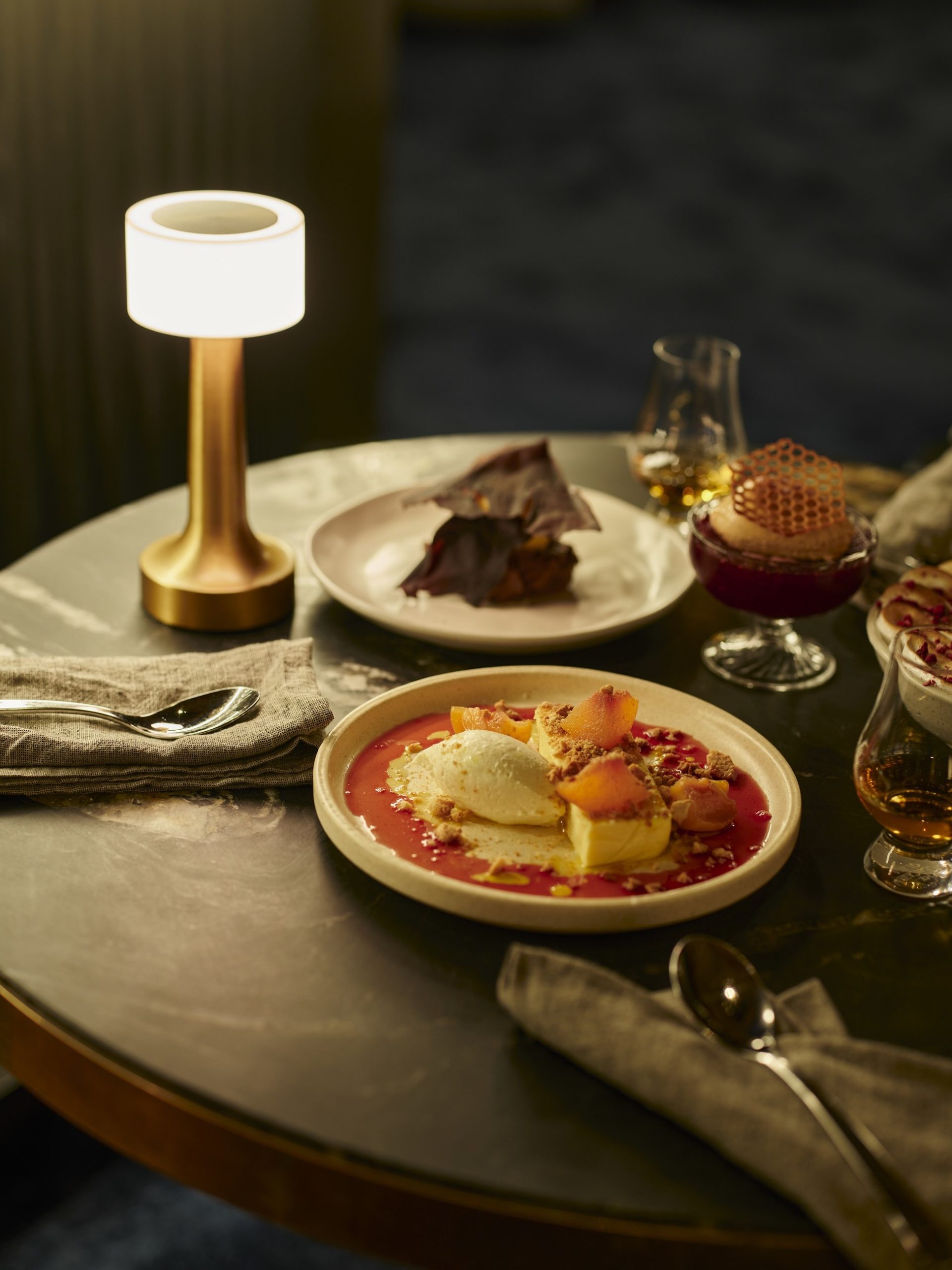 Luxury dessert spread on a round table with brass lamp, berry coulis plate, and whisky glasses.