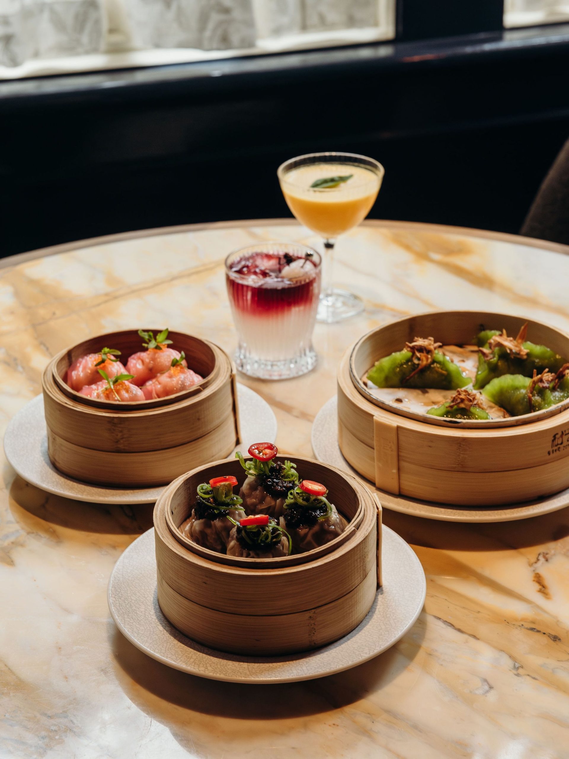 Three bamboo steamers of colourful dim sum beside two cocktails on a marble table.