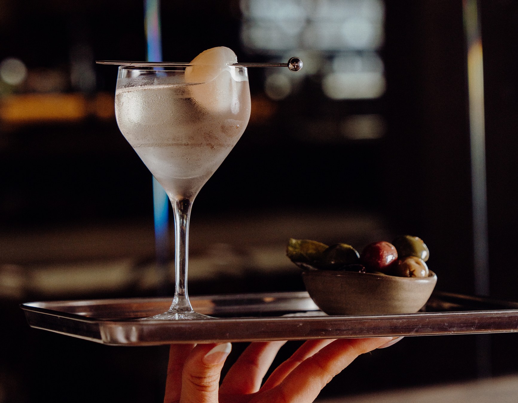 A frosted Gibson martini on a silver tray beside a small bowl of mixed olives, held aloft in a dimly lit bar.