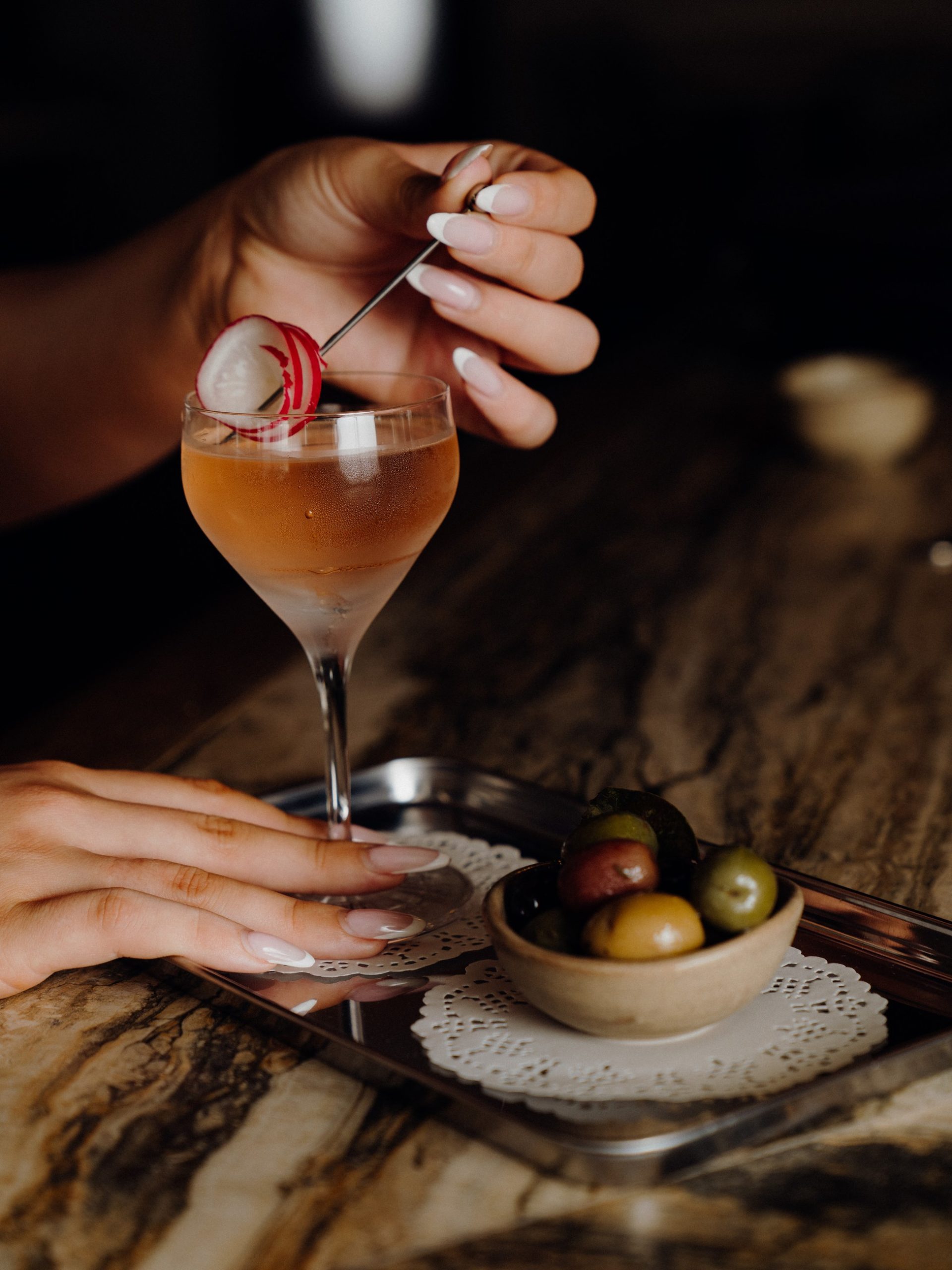 Hands holding a garnished amber cocktail in a coupe glass beside a bowl of olives on a marble bar.