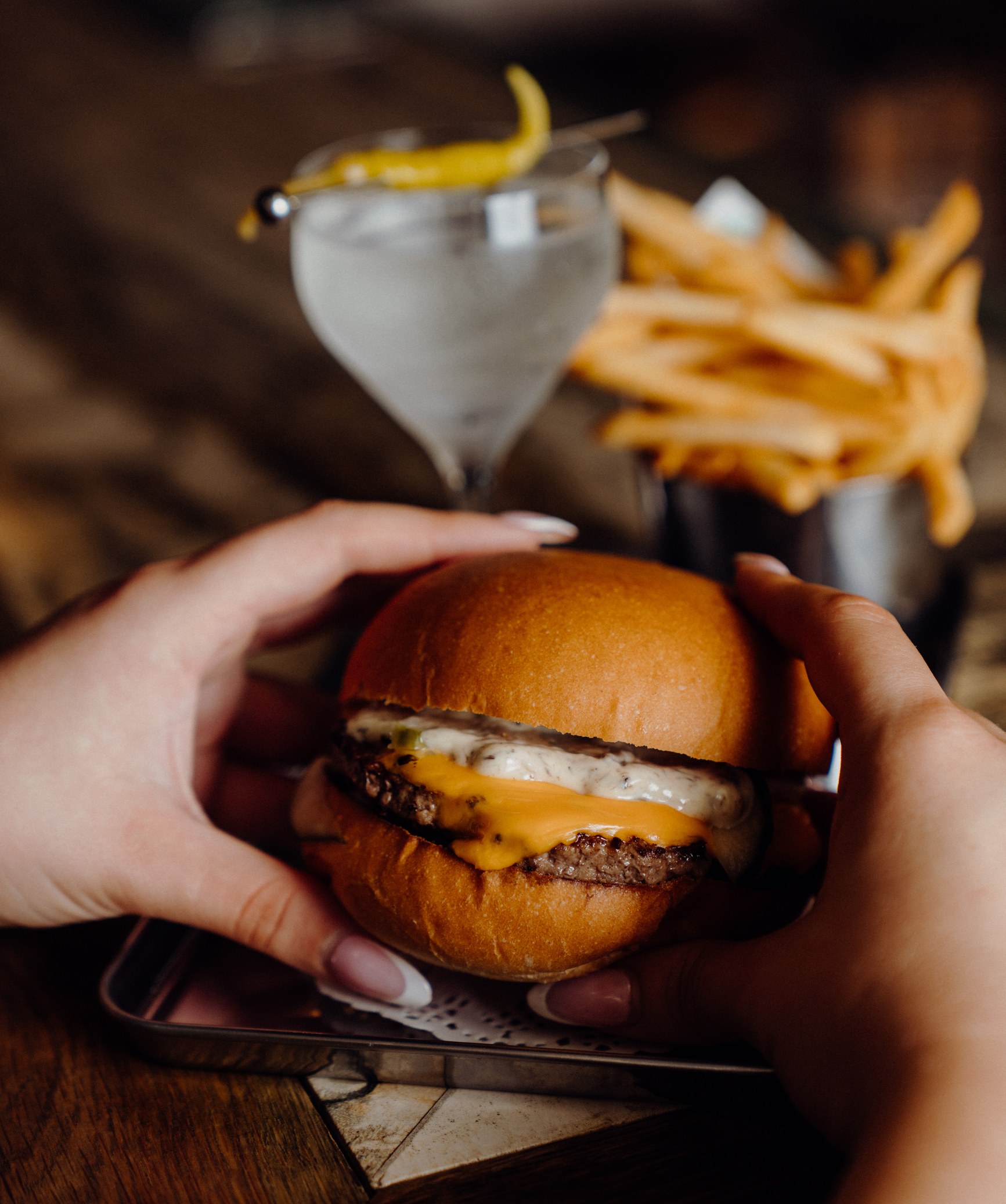 Hands holding a brioche cheeseburger with a dirty martini and fries in the background.