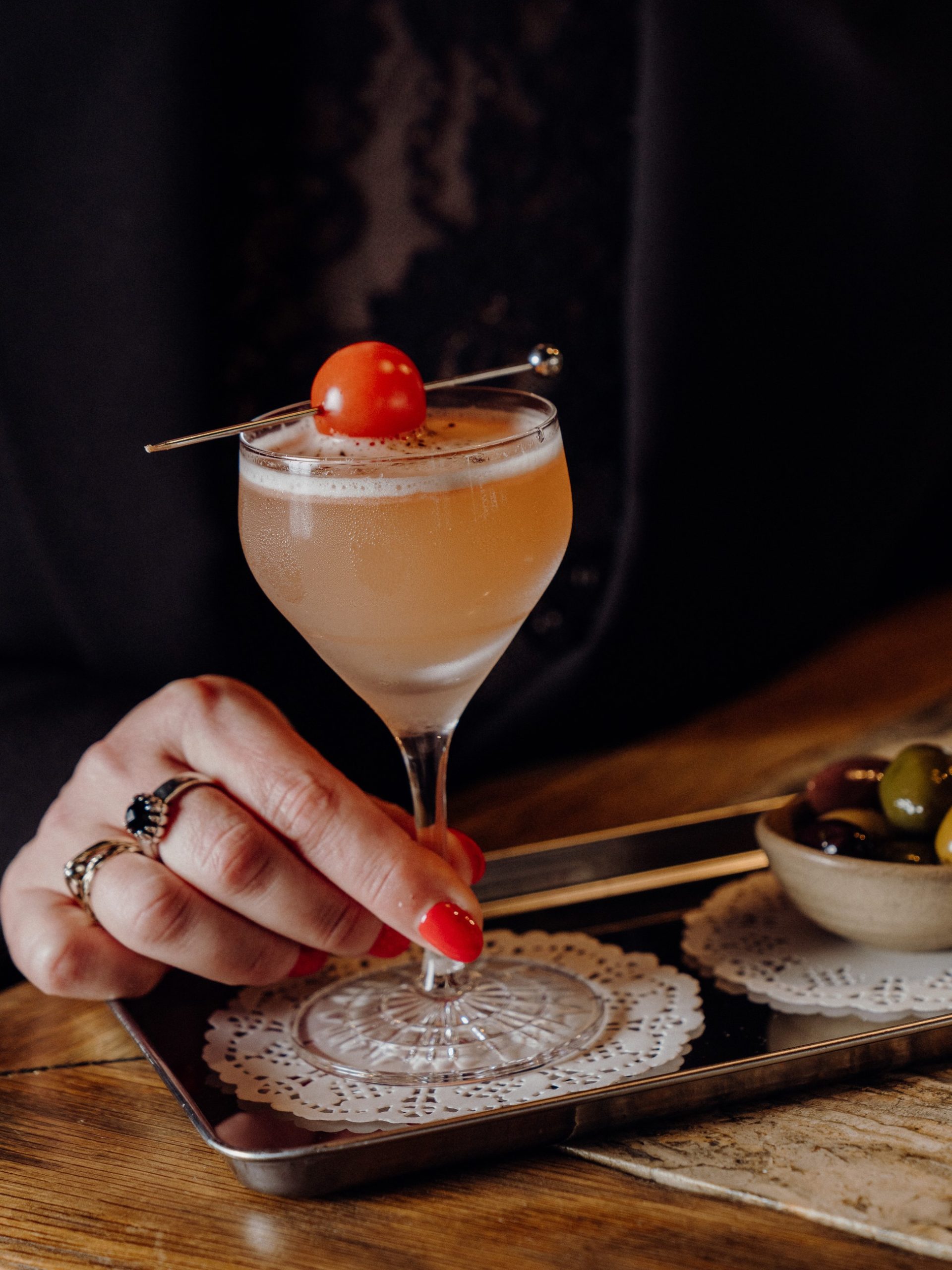 A hand with red nails holds a blush martini in a crystal coupe garnished with a cherry tomato.