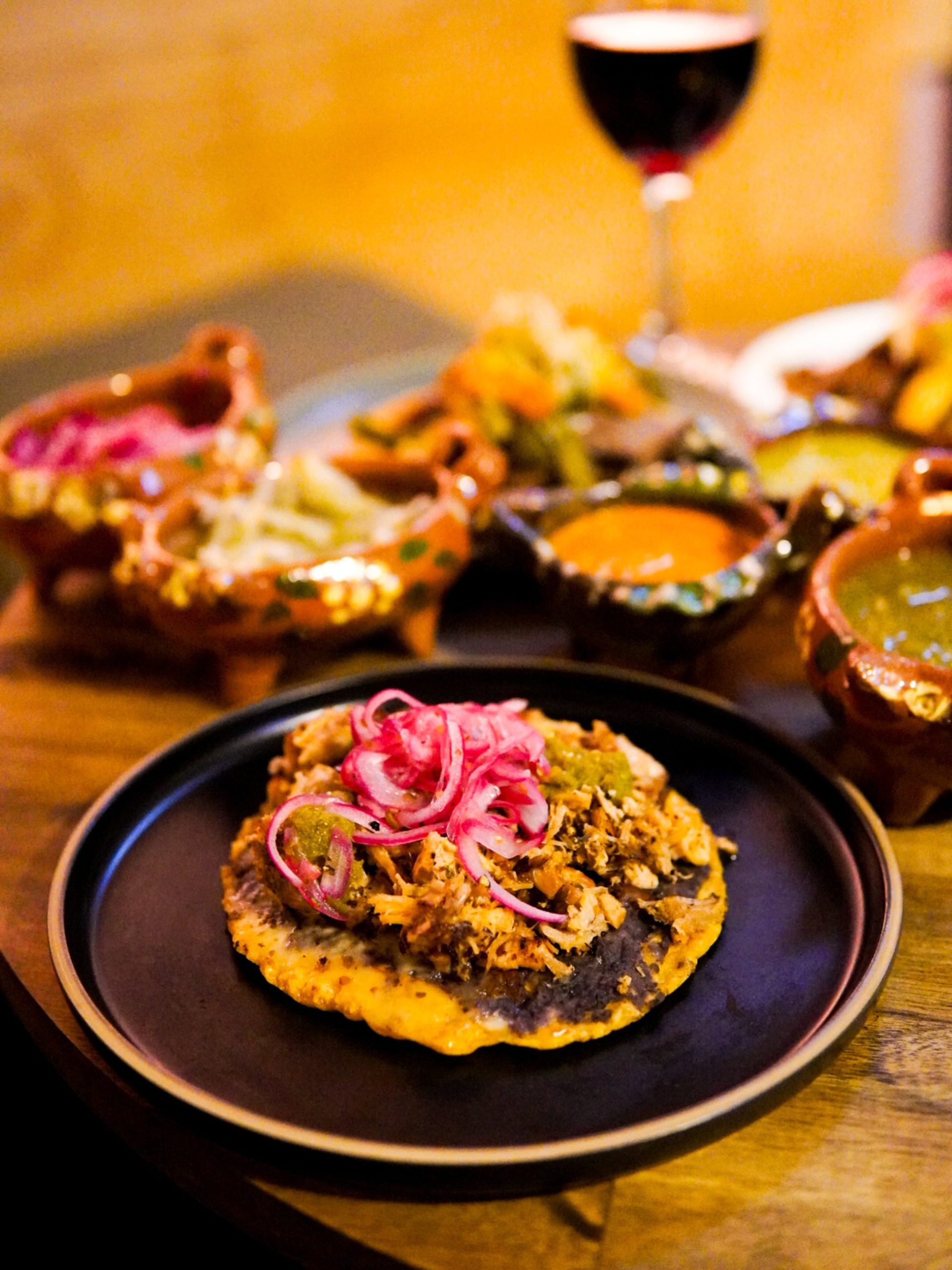 A tostada with braised meat and pickled red onions on a black plate, salsas and red wine behind.