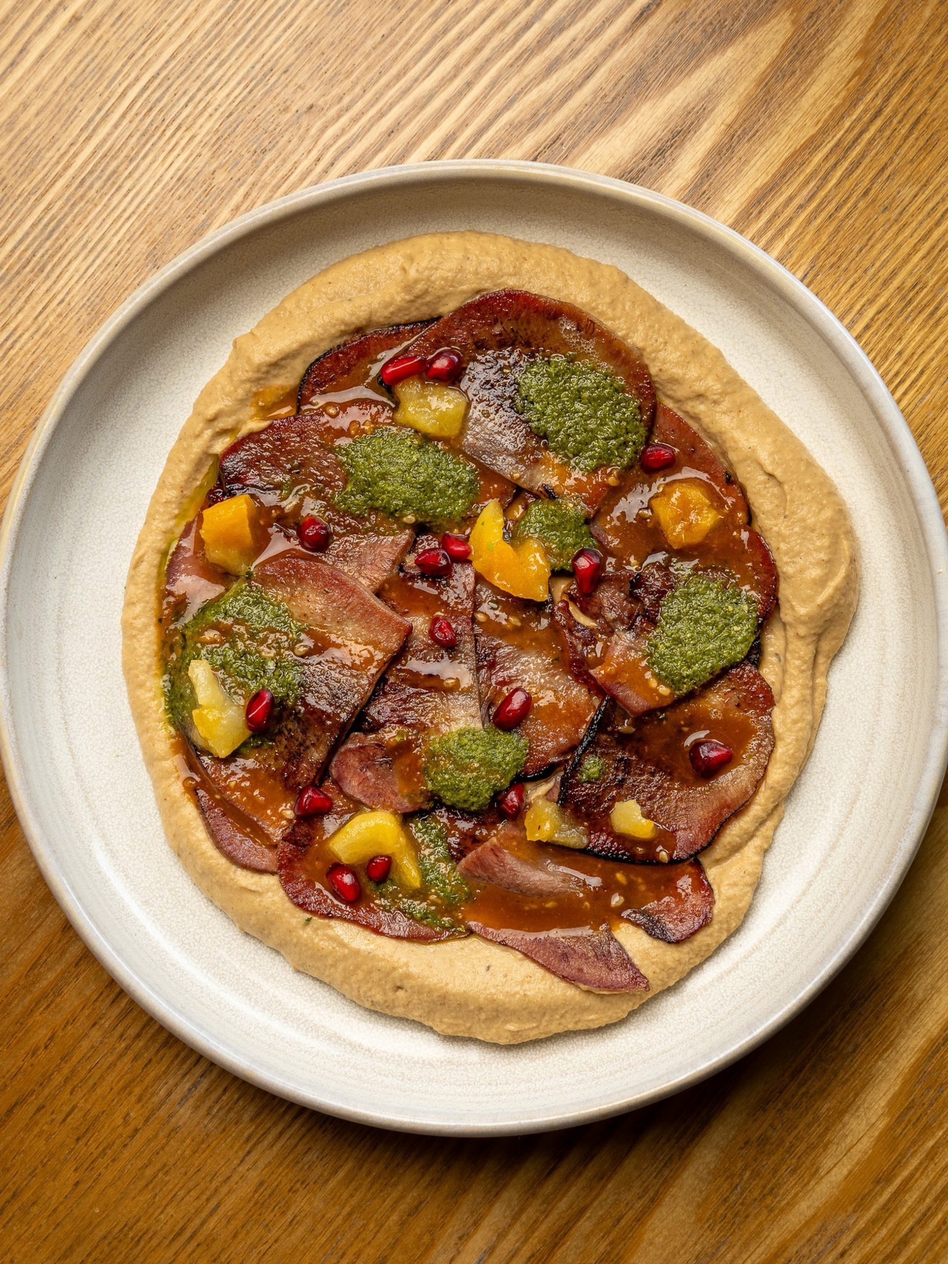 Overhead view of a flatbread with charred meat, hummus, herb sauce, pomegranate seeds, and diced fruit on a ceramic plate.