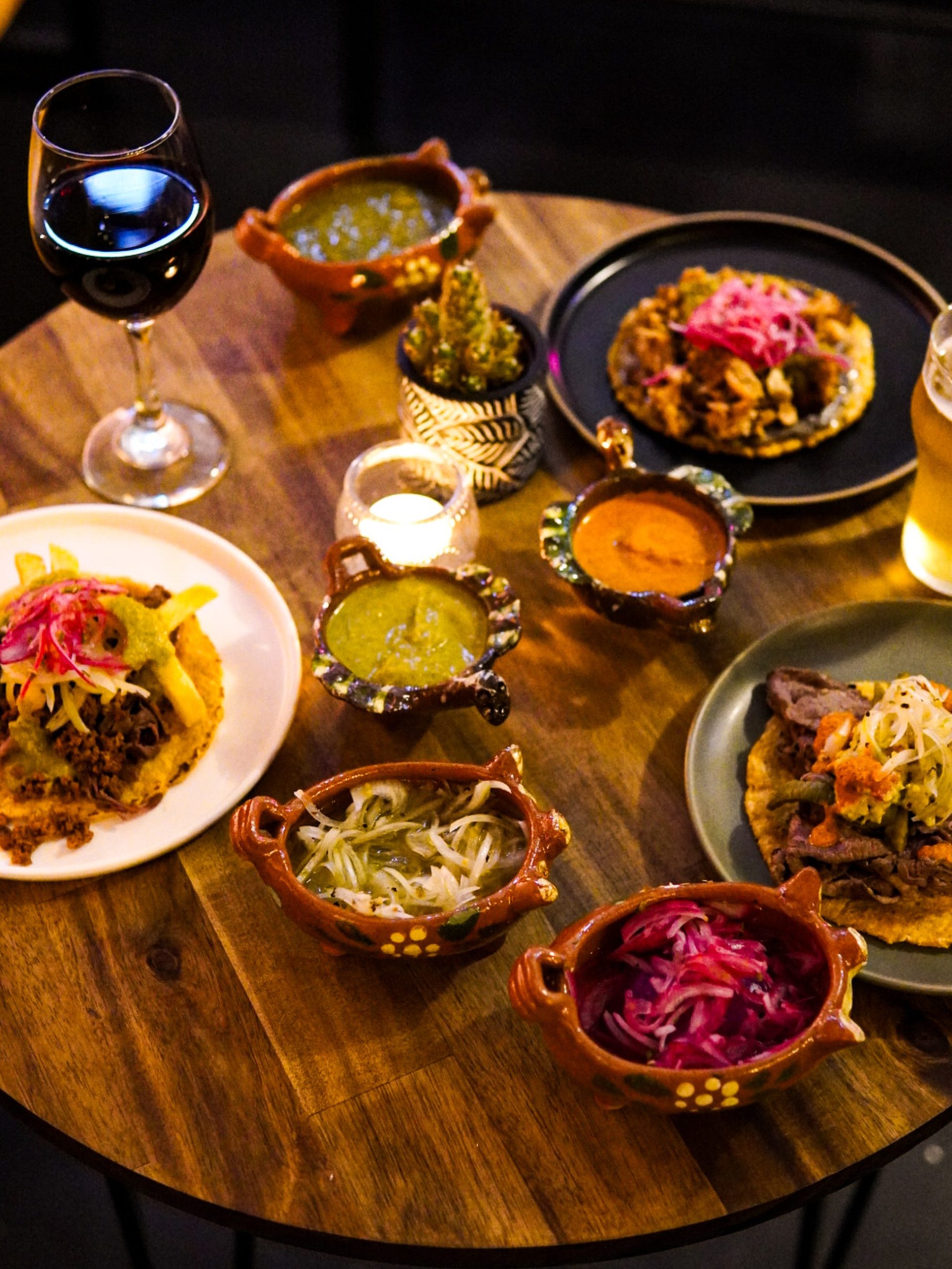 Overhead view of a Mexican restaurant table spread with tostadas, salsas in terracotta bowls, wine and beer.