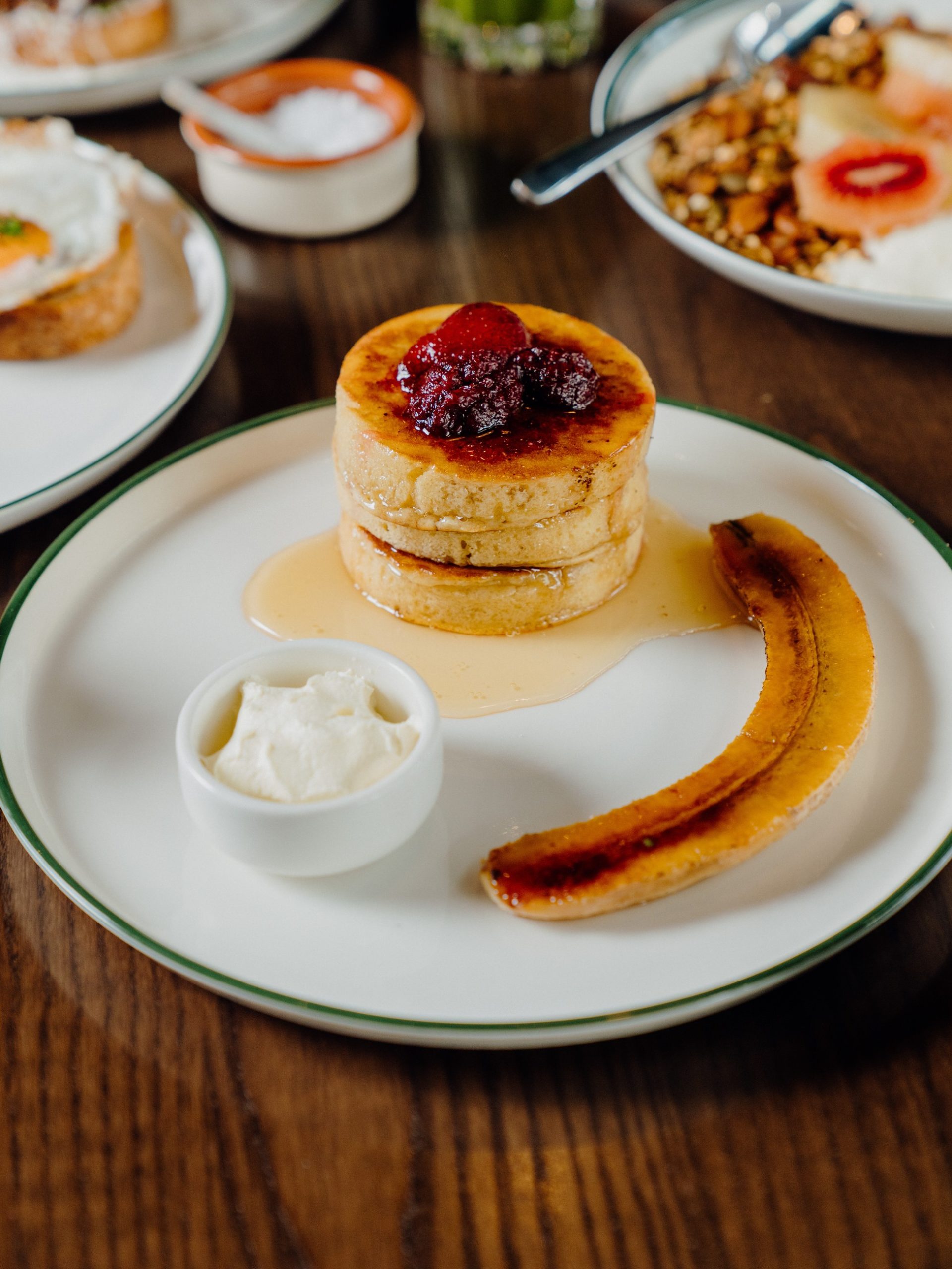 Stack of golden pancakes with caramelised berries, banana, and whipped cream on a white plate.