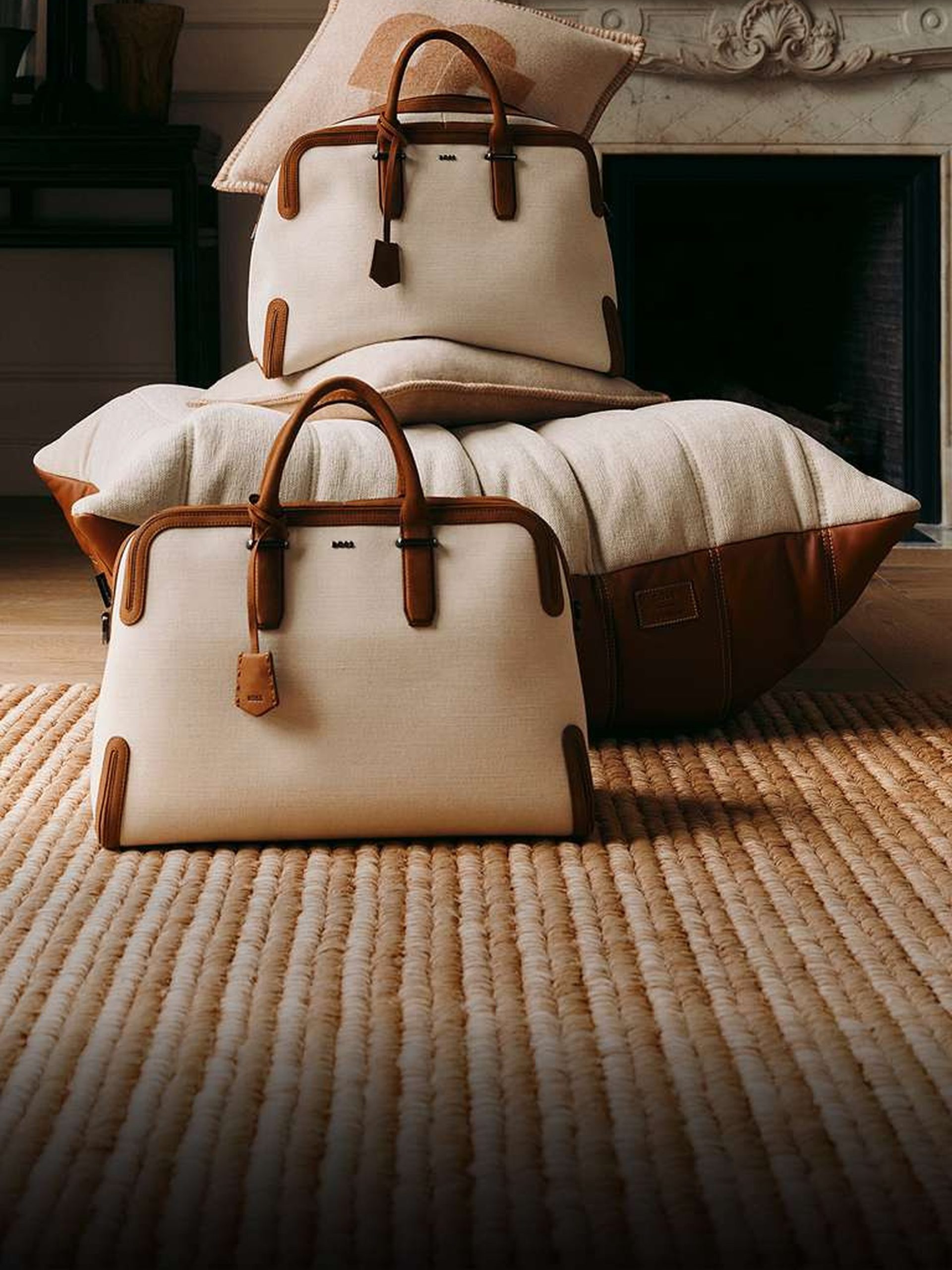 Two cream canvas and tan leather structured handbags styled on cushions before a marble fireplace.