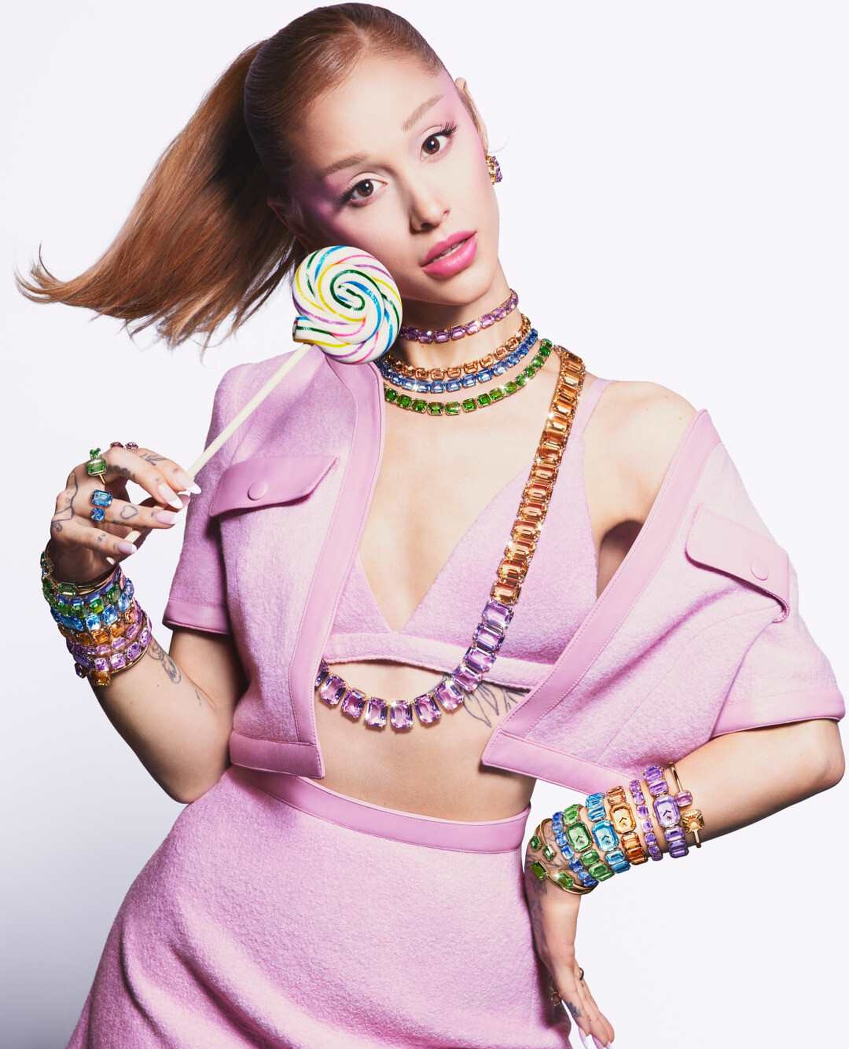 Woman in pink outfit wearing layered Swarovski crystal jewellery holding a rainbow lollipop.