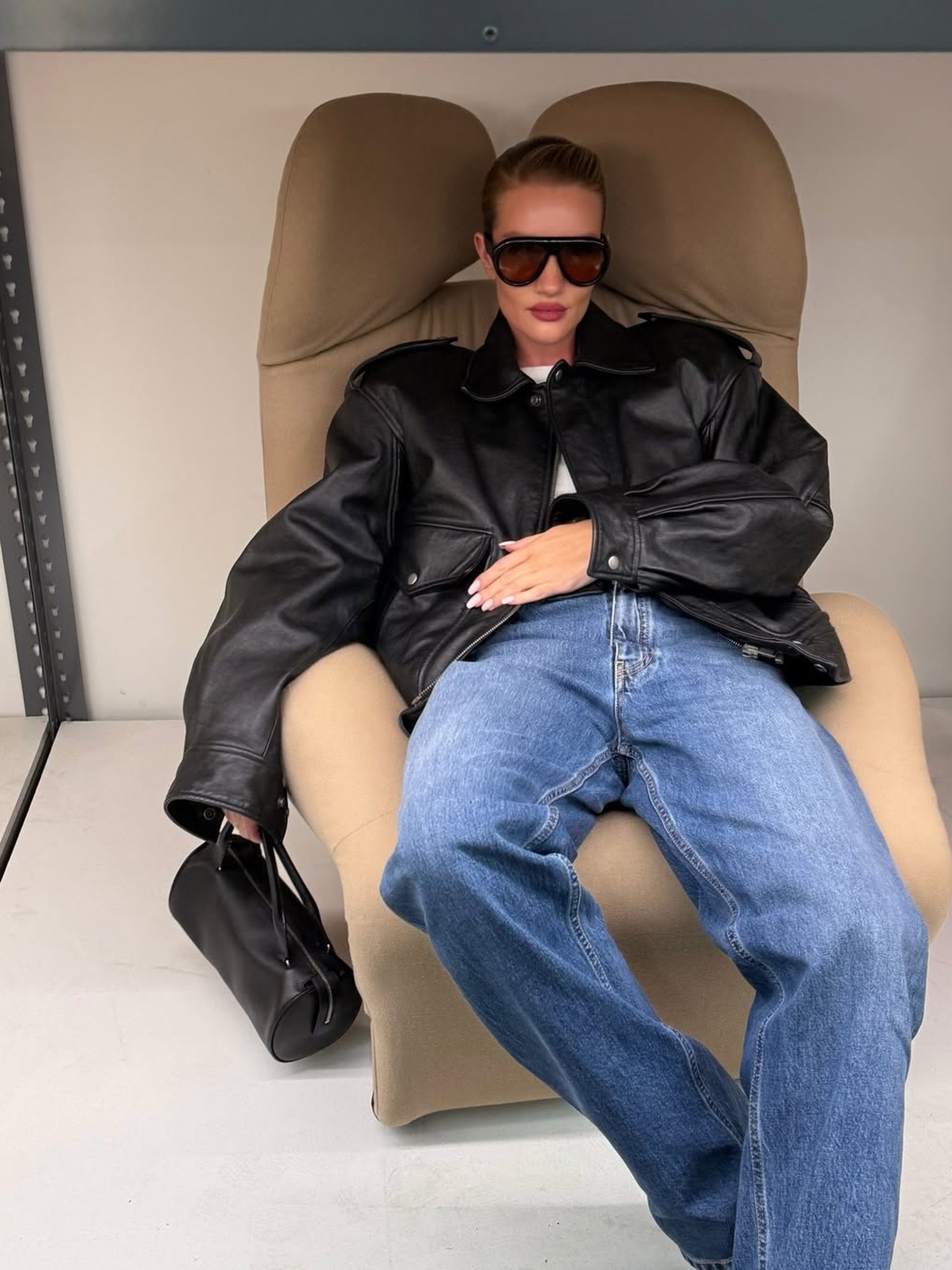 Woman in oversized black leather jacket and jeans reclines in beige lounge chair holding black handbag.