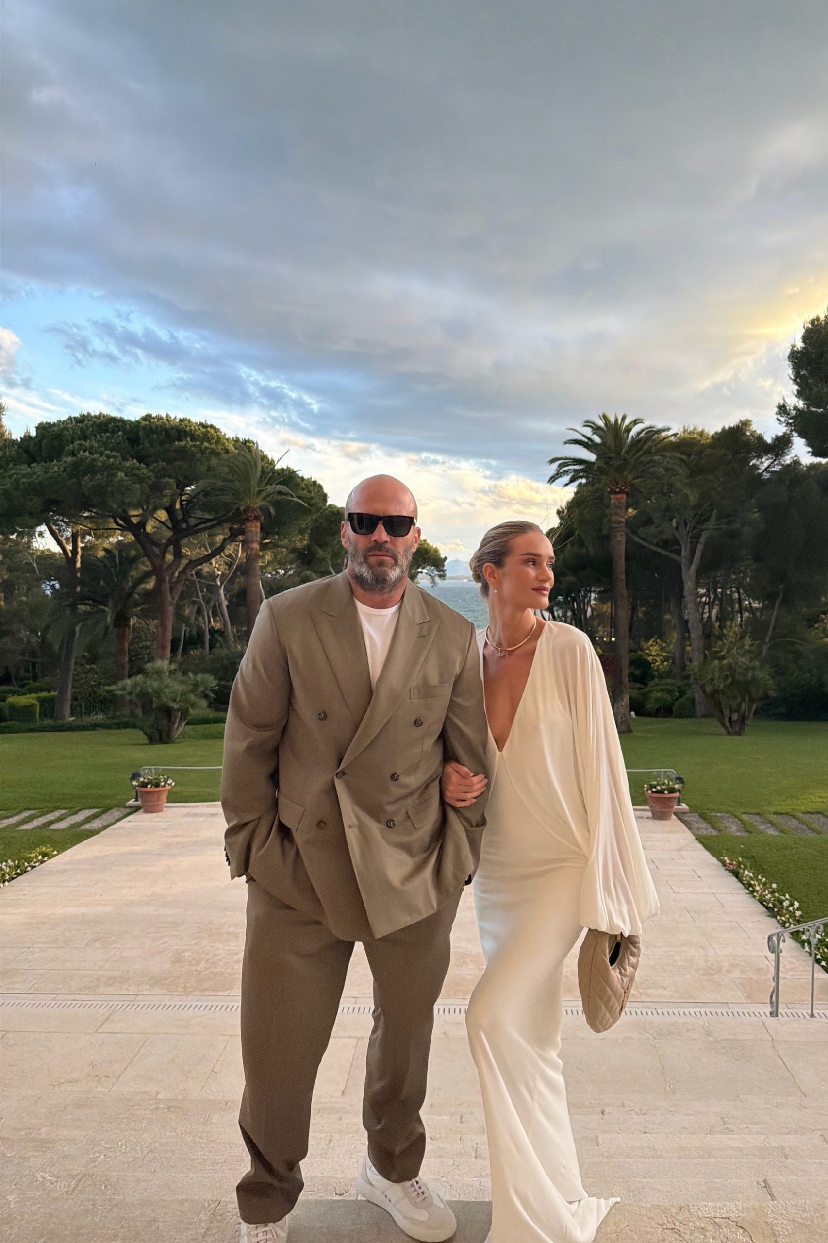 A couple dressed in elegant neutral tones posing on a terrace with Mediterranean gardens behind them.