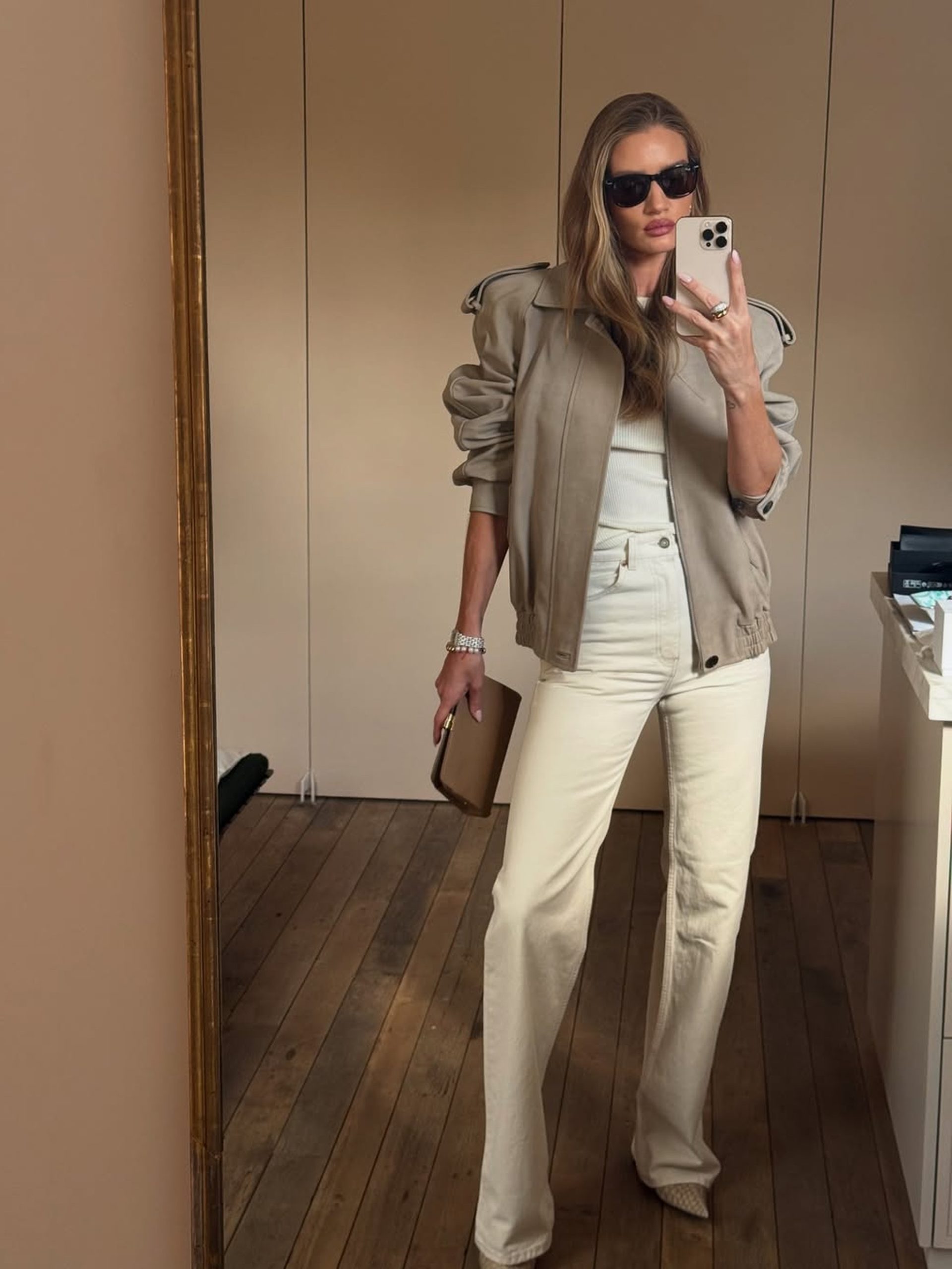 Woman in tan jacket, cream wide-leg jeans and sunglasses takes a mirror selfie holding a clutch.