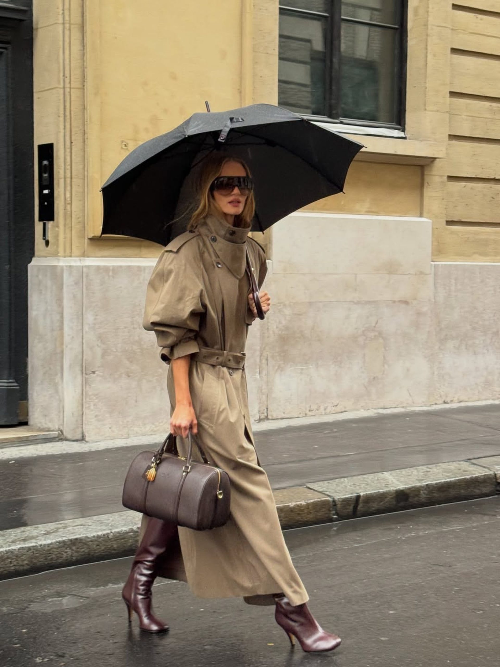 Woman in belted khaki trench coat and burgundy boots walking in rain on Parisian street.