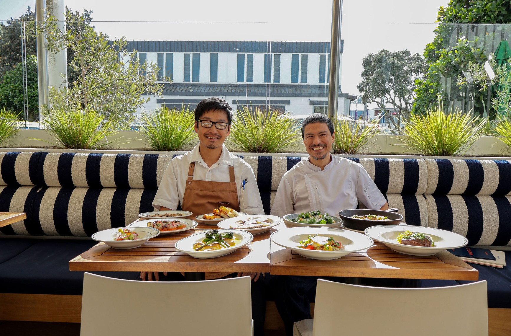 Two smiling chefs seated behind a full spread of plated dishes at a modern Auckland restaurant.