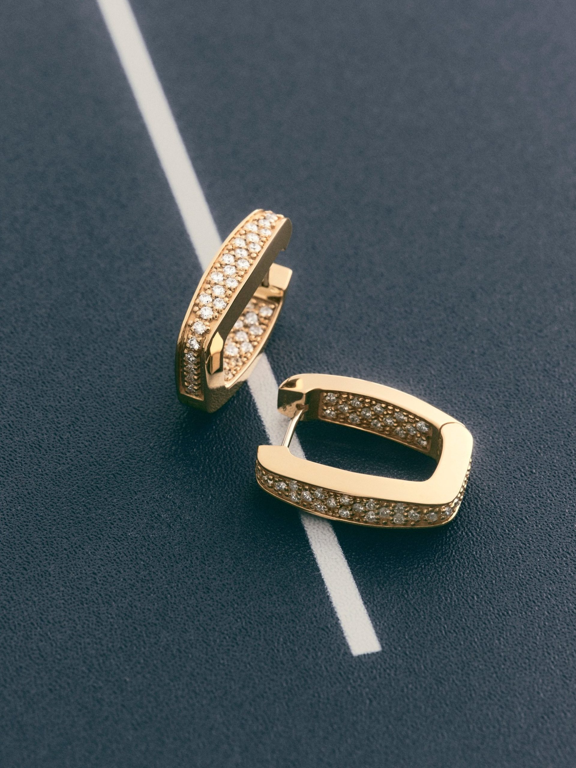 Two rectangular gold pavé diamond hoop earrings placed on a dark navy surface with a white line.