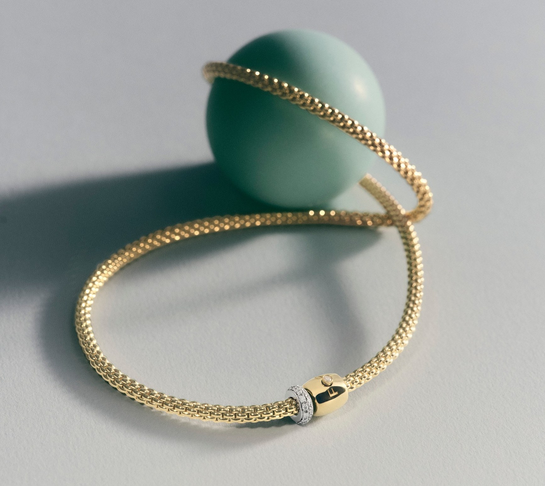 Gold mesh bracelet with pavé diamond clasp draped around a mint green sphere on grey background.