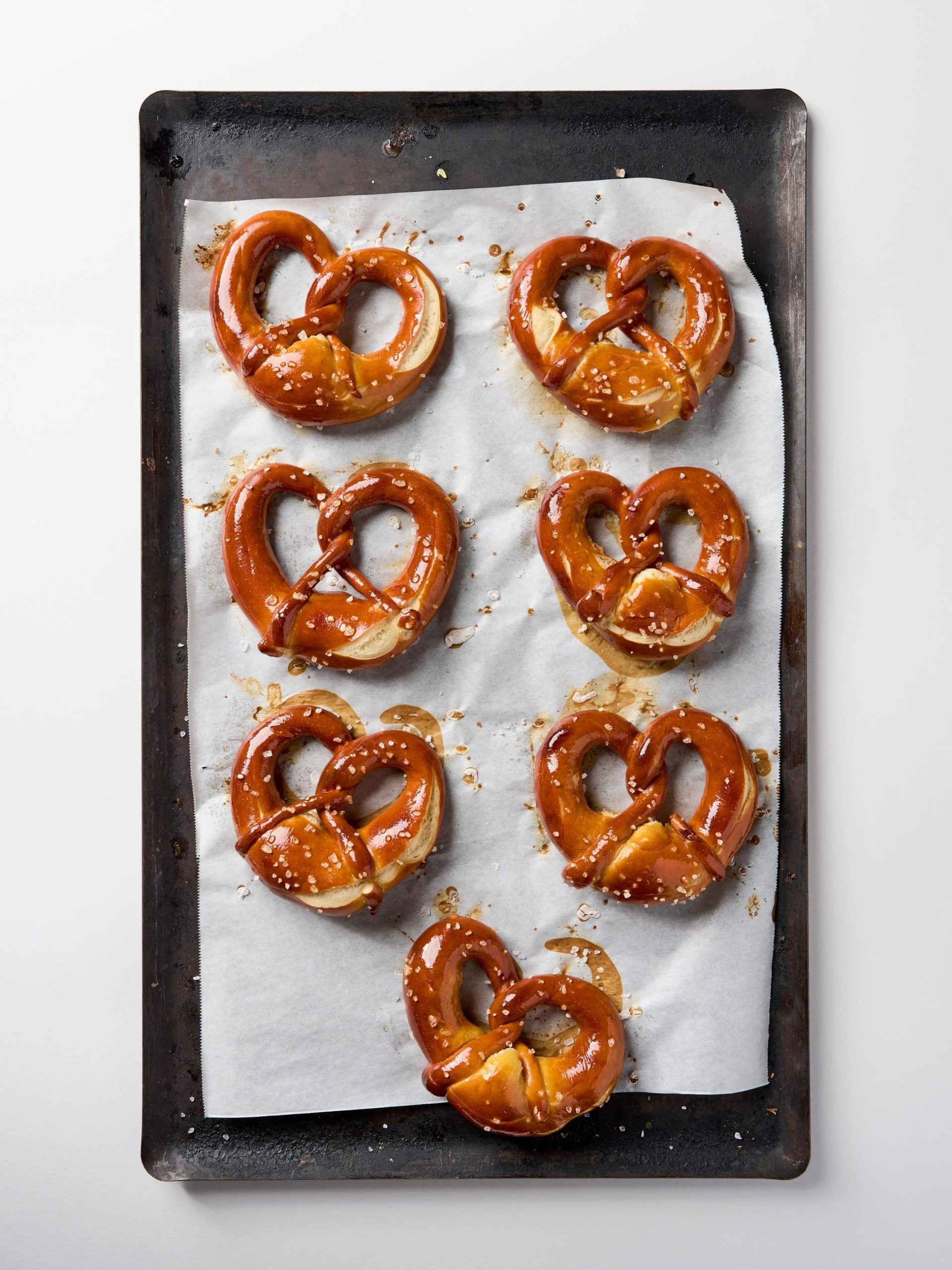 Seven glossy baked soft pretzels with coarse salt on parchment-lined dark baking tray, overhead view.