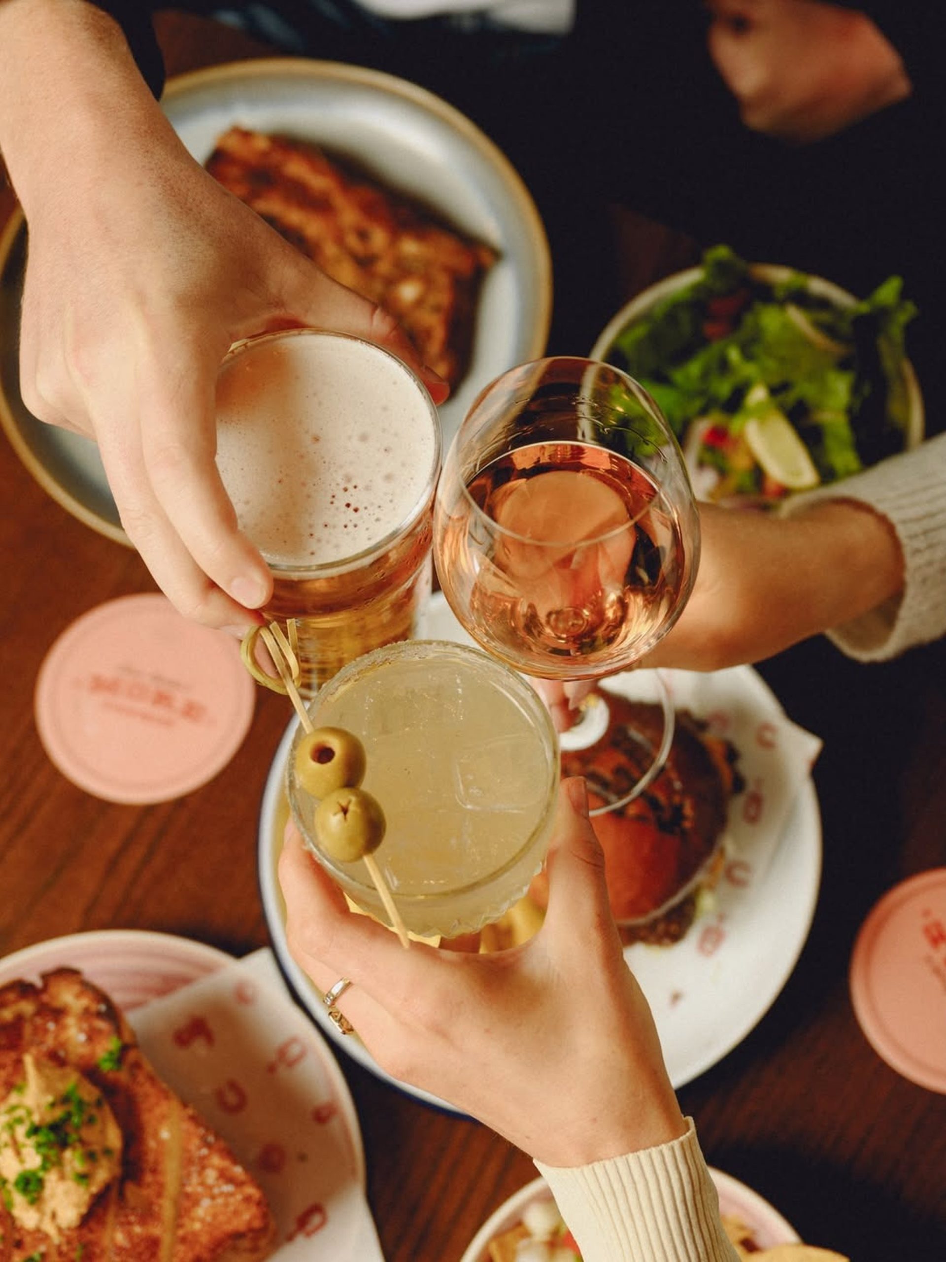 Three hands toasting with beer, rosé, and an olive cocktail over a shared restaurant table.